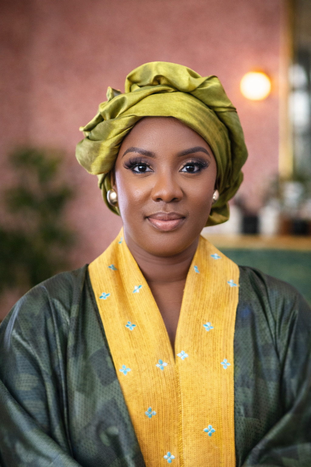 A woman wearing traditional African attire, including a green headwrap and a matching gown with yellow and blue embroidery, poses indoors with a pinkish wall background and warm lighting.