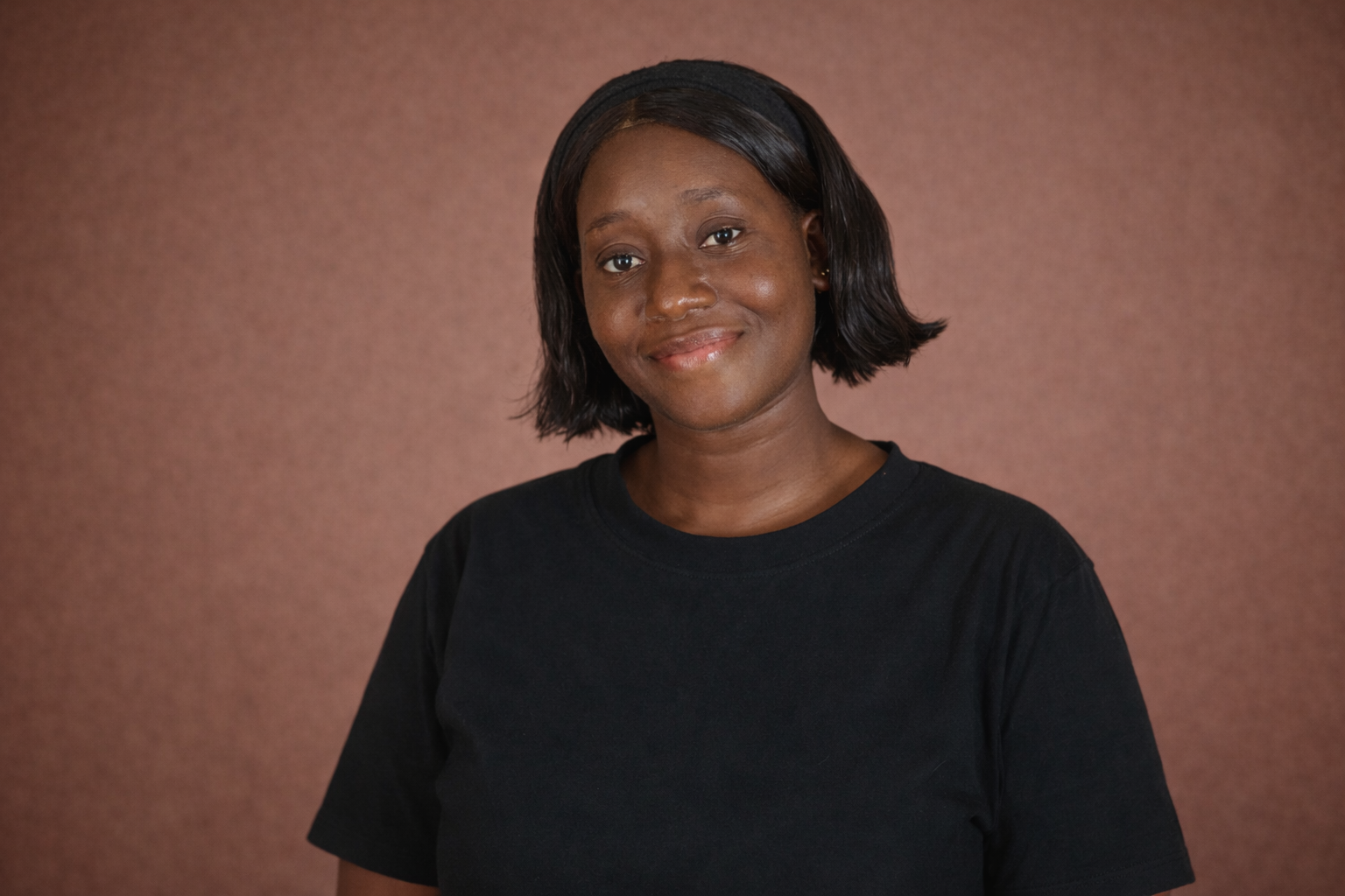 A young woman with medium skin tone and short black hair, wearing a black t-shirt, smiling against a brown background.