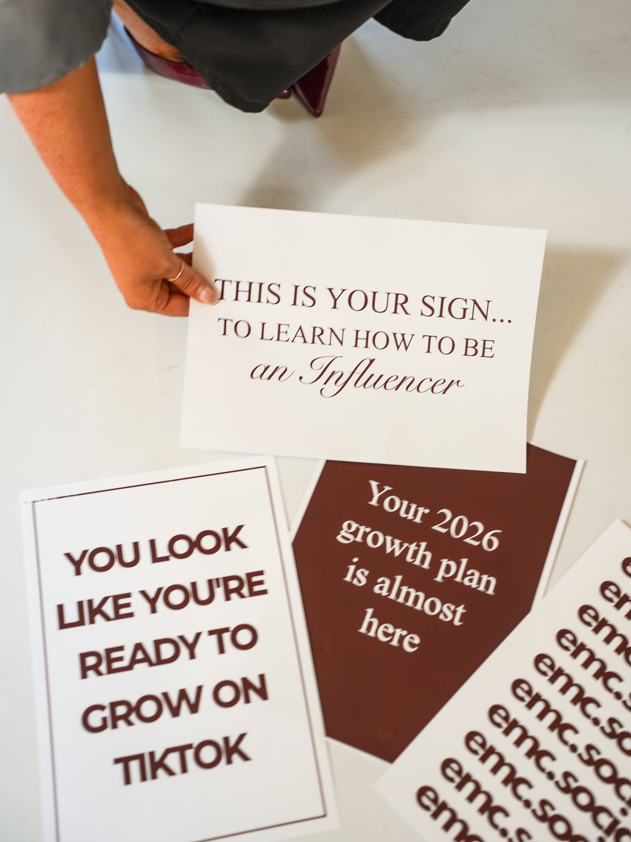 A person holding a white card with the text 'This is your sign... to learn how to be an Influencer.' Other similar cards with motivational phrases about growth and achievement are visible on the table.