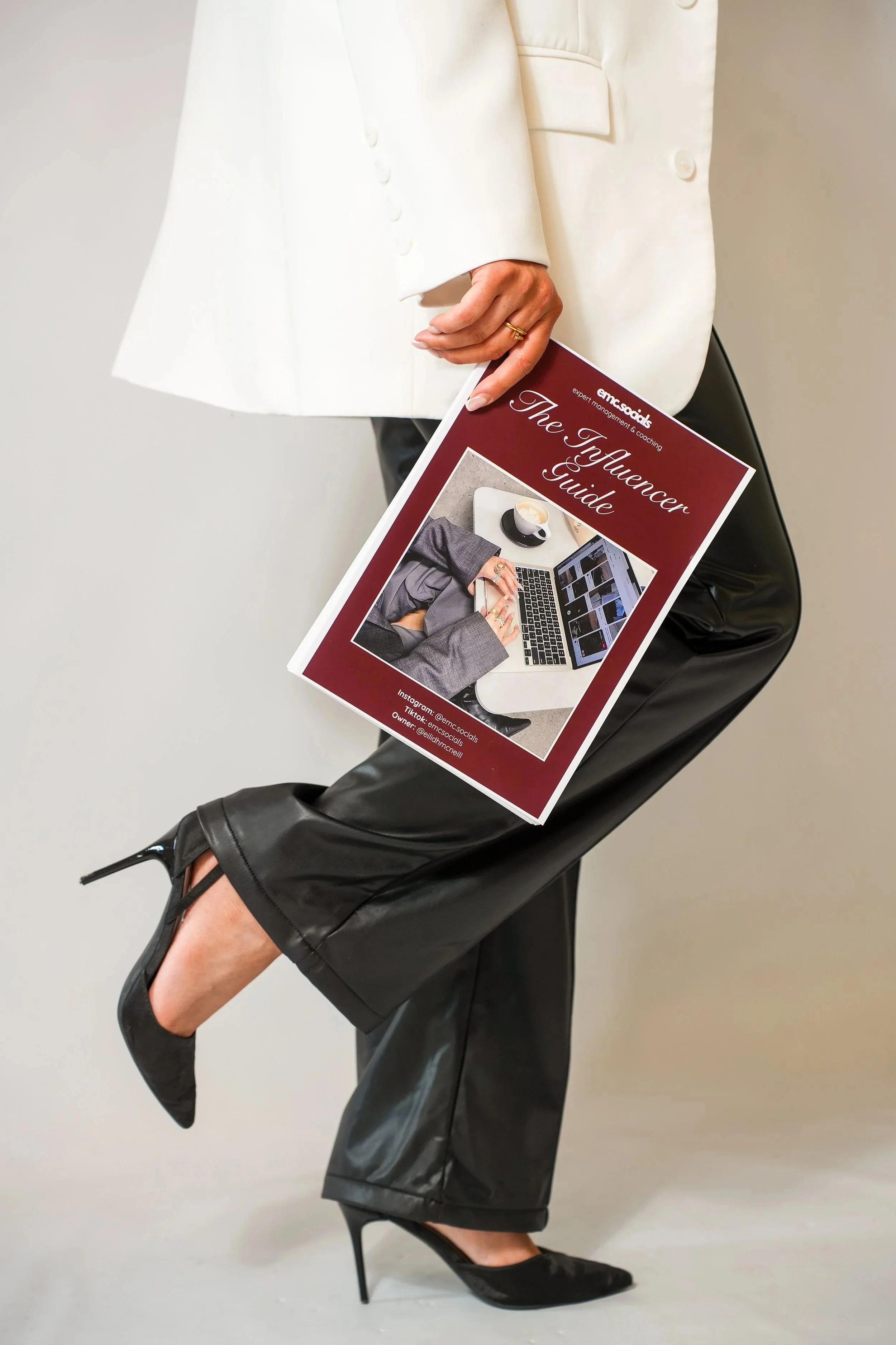 A woman in black high heels, a black leather skirt, and a white blazer sitting on a chair, holding a magazine titled 'The Influencer Guide' with a photo of a person using a laptop on the cover.