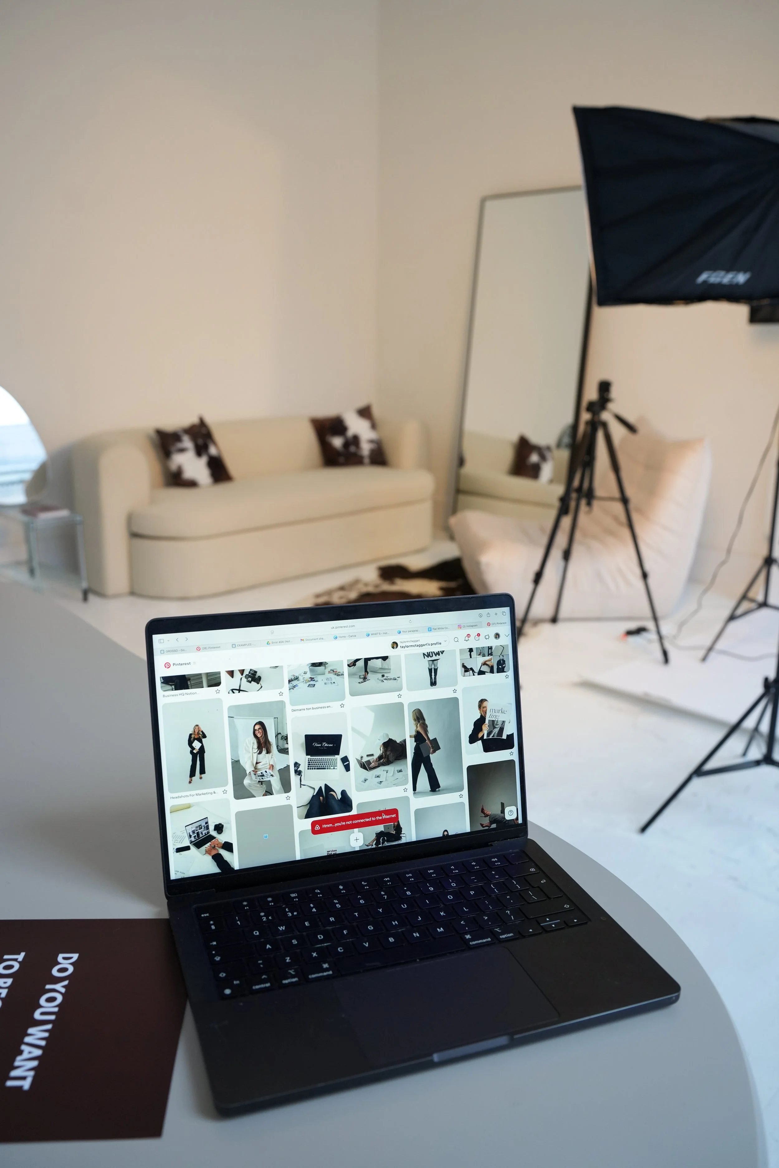 Photo of a photography studio setup showing a white couch with black and white cow-patterned pillows, a large mirror, studio lighting with a softbox, a tripod, and a laptop displaying a Pinterest page with images of women in stylish outfits.