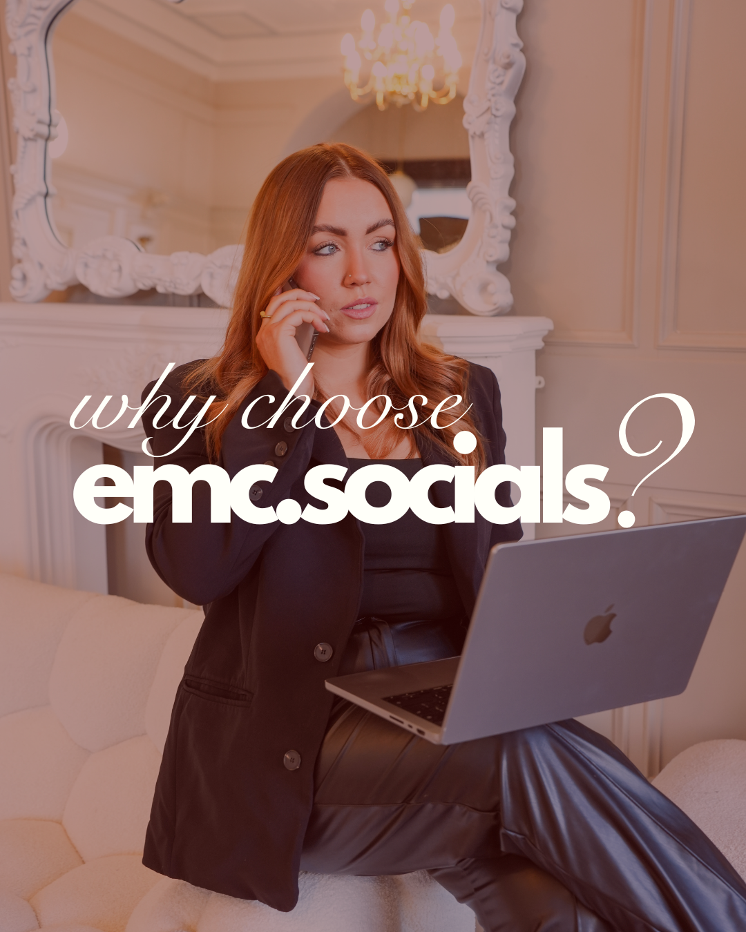 A woman with red hair, wearing a black blazer and leather pants, sitting on a cream-colored couch with a laptop on her lap and talking on a phone. There is a white ornate mirror and chandelier in the background.