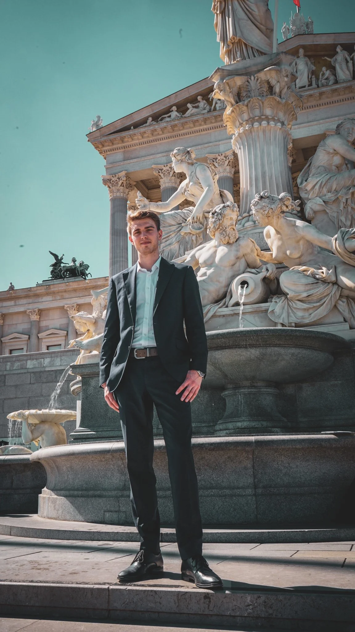 A young man in a black suit and white shirt stands in front of a neoclassical marble fountain with statues and figures, with a building in the background and a clear sky.