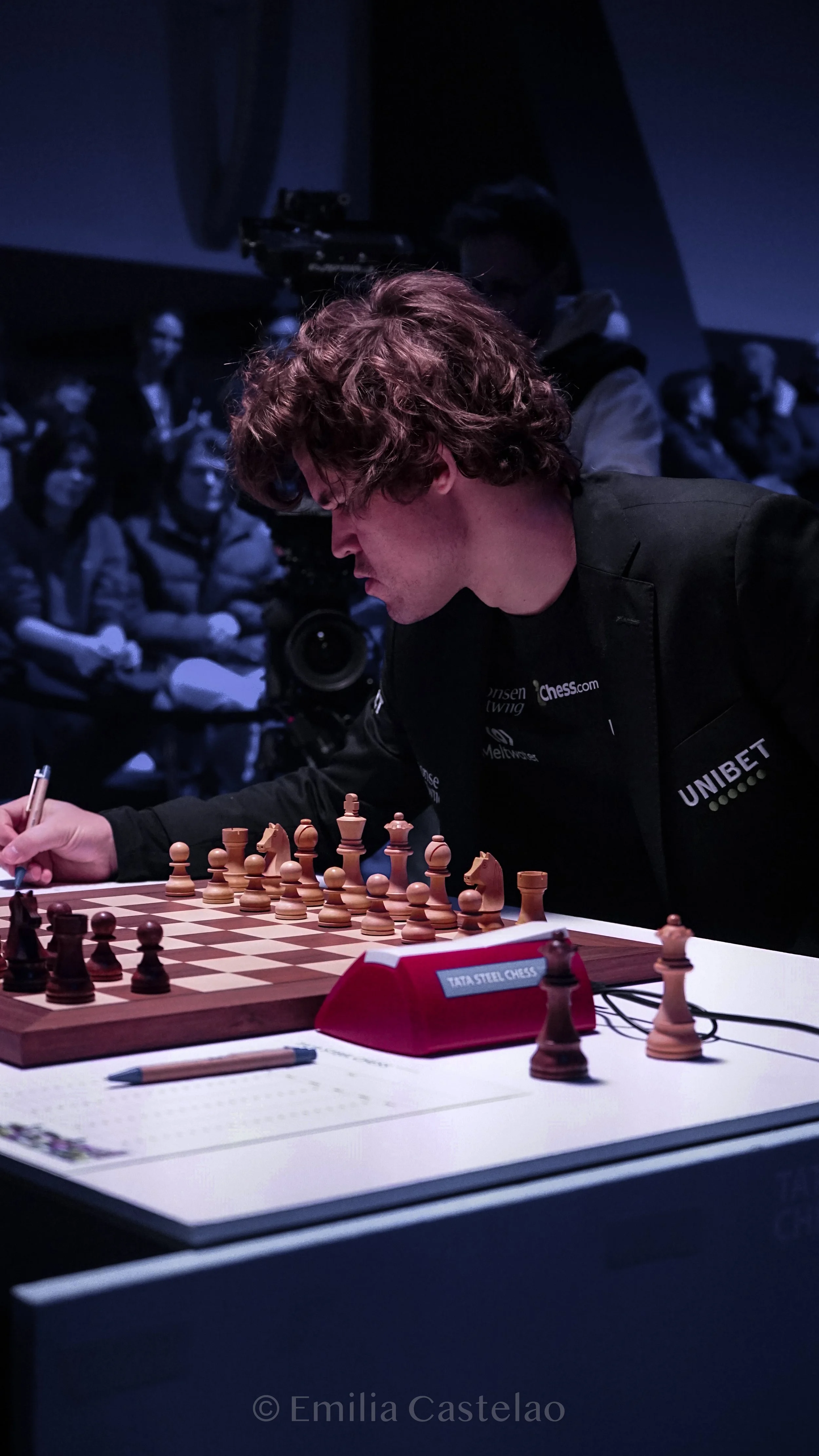 A young man with curly hair playing chess during a tournament, with spectators and a cameraman in the background.