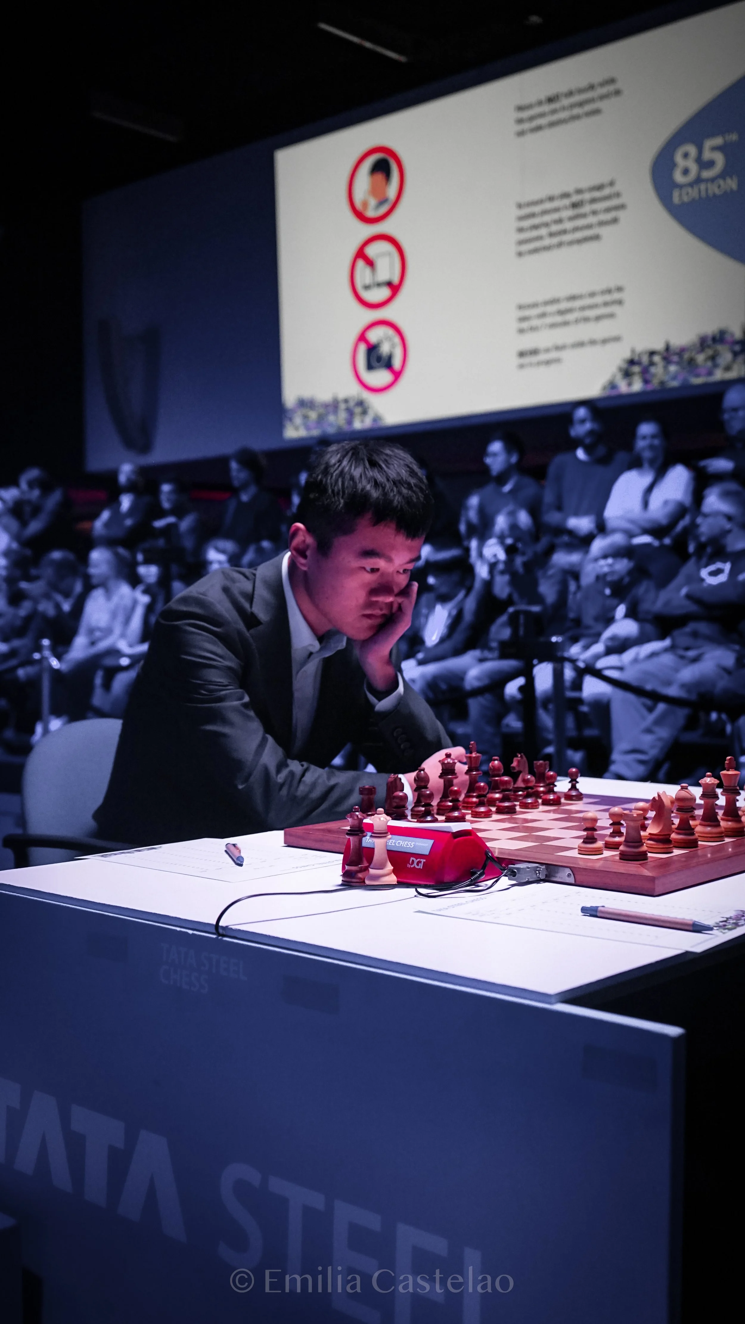A man playing chess in a tournament, seated at a table with a chess clock and notes, with a large audience watching in the background and a screen displaying rules and information.