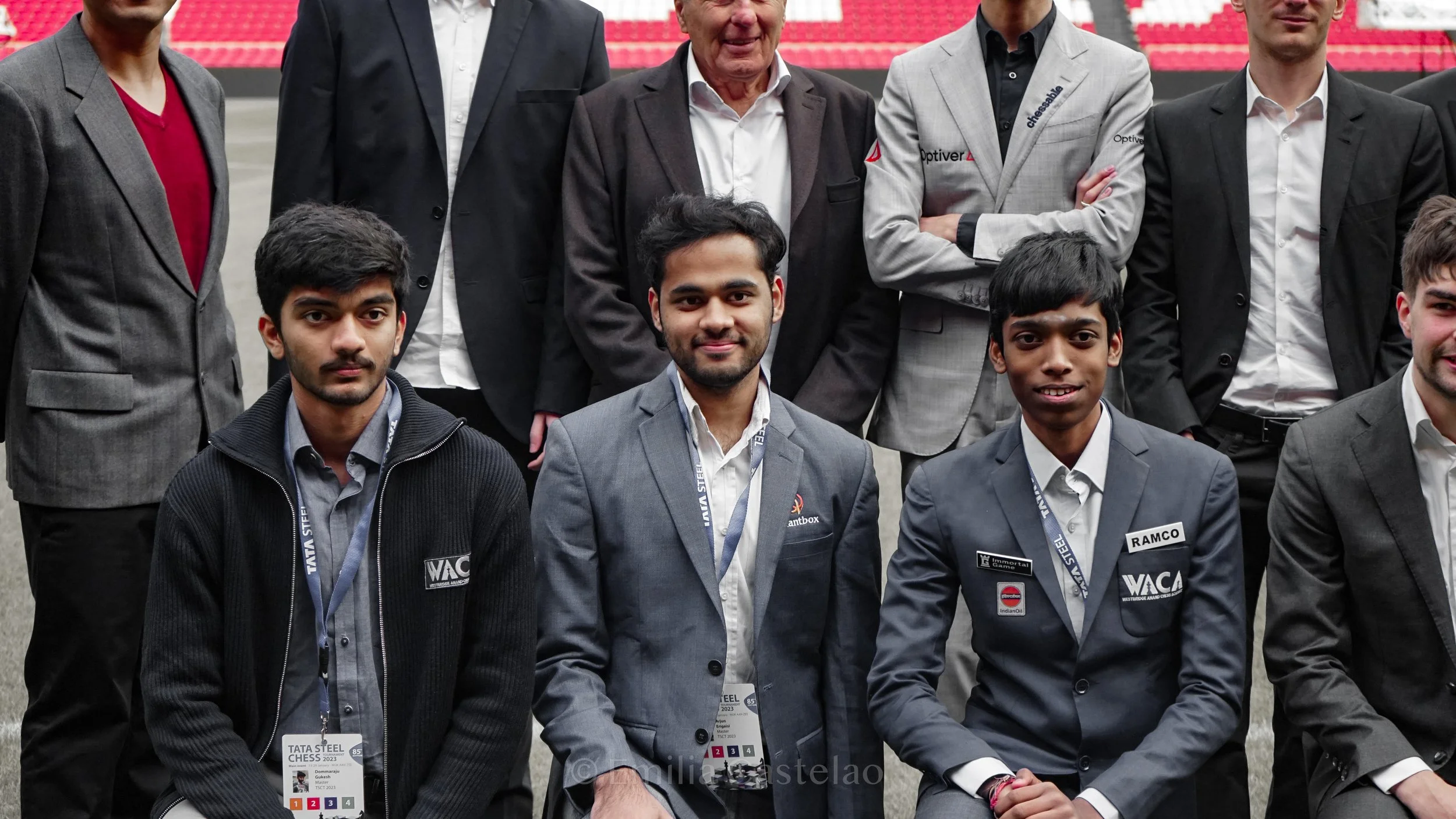 Group of young men and older gentlemen in formal attire posing for a photo on a chess tournament stage, with red seats in the background.