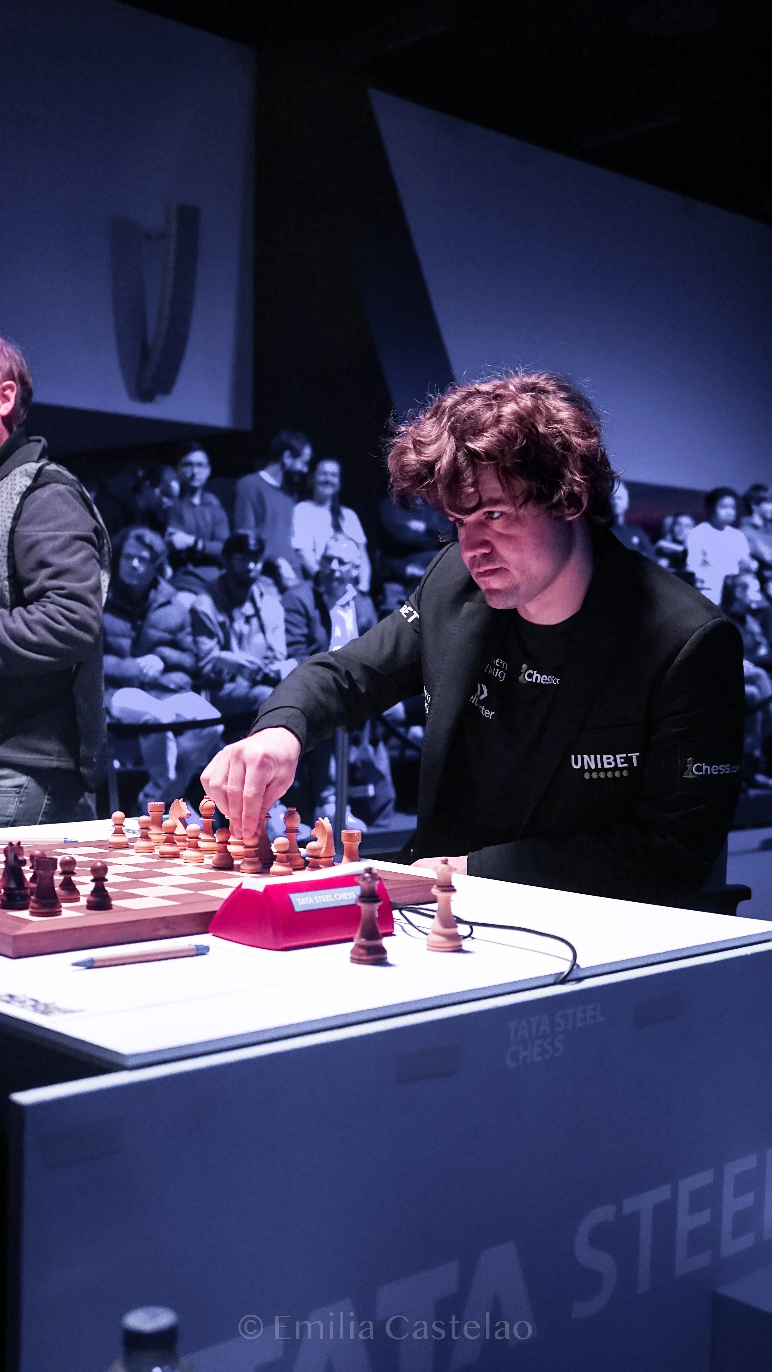 A young man with curly hair playing chess at a tournament table, with a crowd of spectators observing in the background.