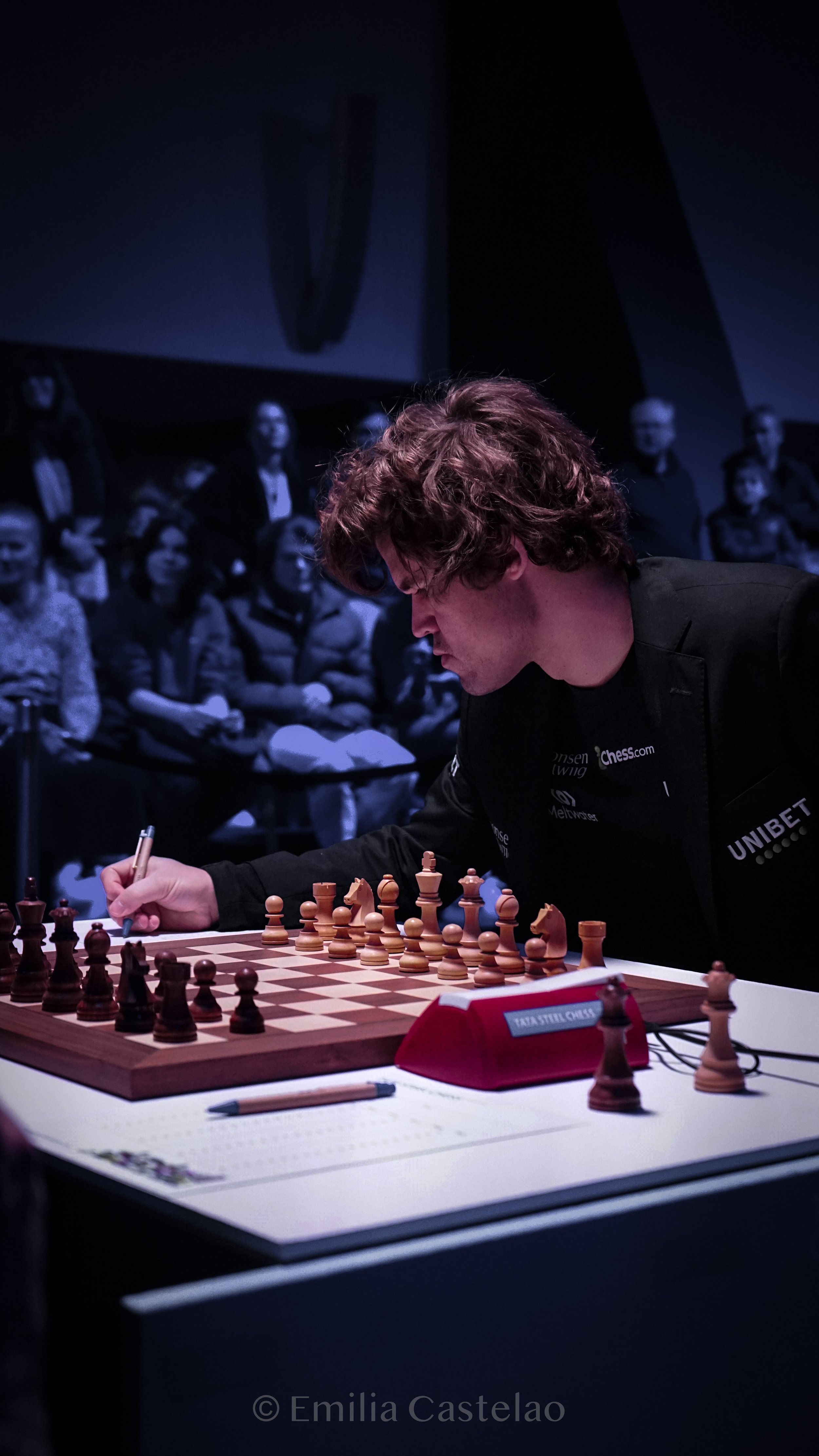 A man playing chess during a tournament with an audience in the background. The player is making a move at the chessboard with chess pieces set up for a game.