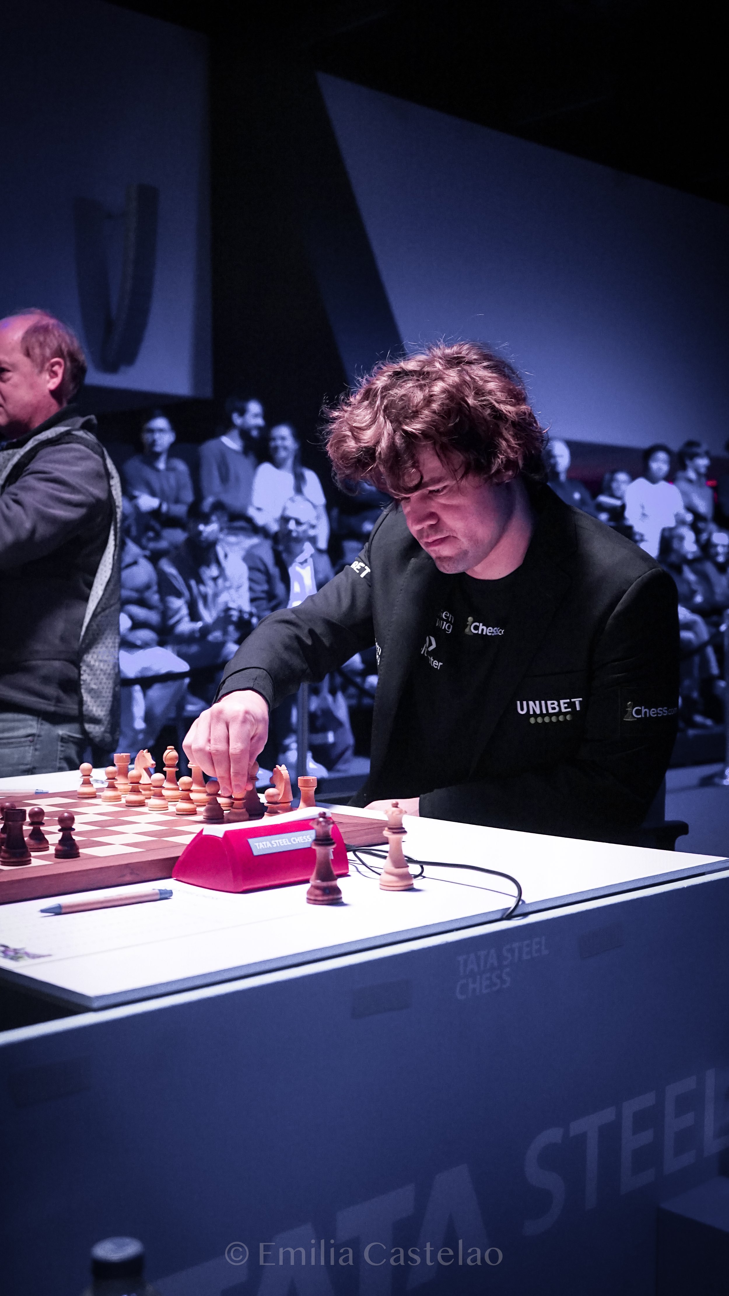 A man with curly hair concentrating on a chess game at a tournament, with spectators in the background. The chessboard is on a table with a chess clock and a pen nearby.