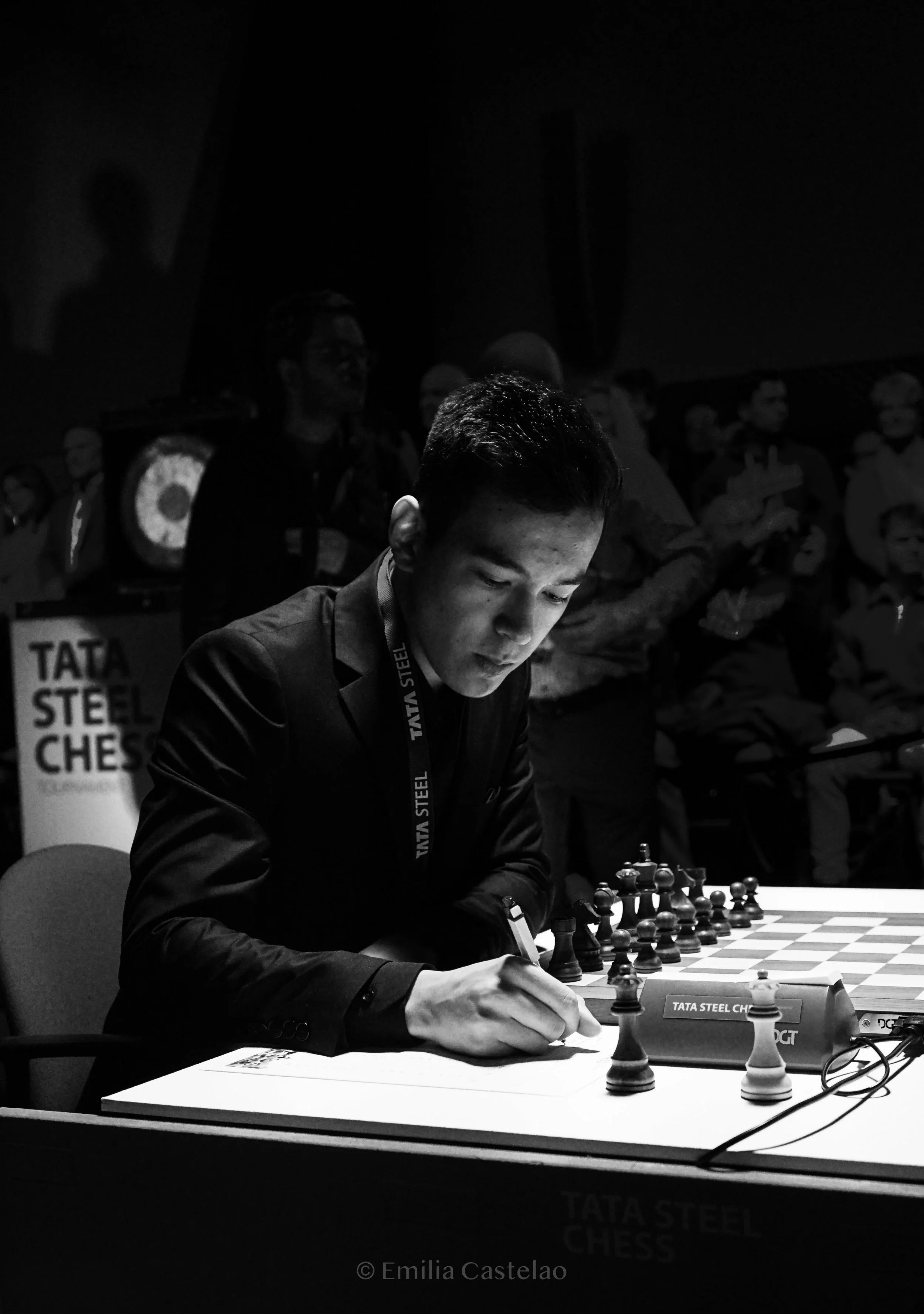 A young man in a suit playing chess during a tournament, with several onlookers in the background.