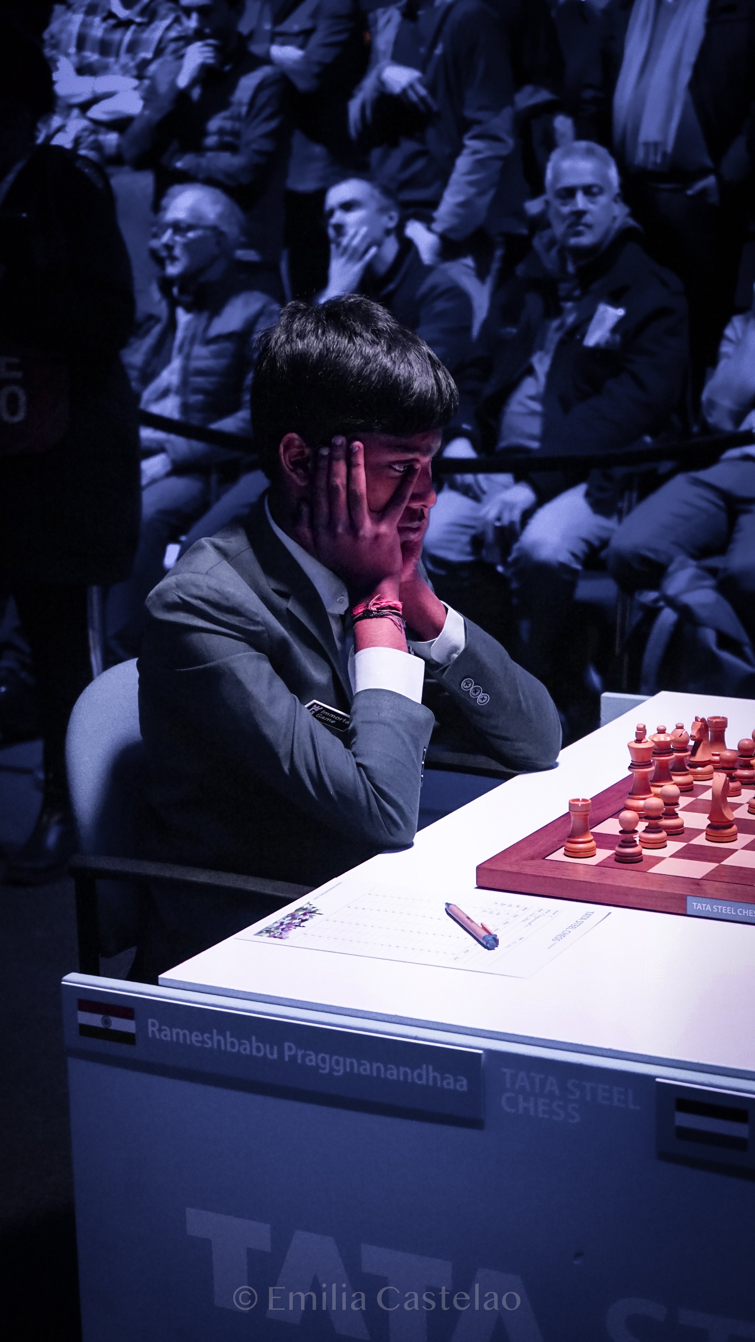 Young chess player in a suit, with hands on face, competing in a chess tournament, with spectators watching in the background.