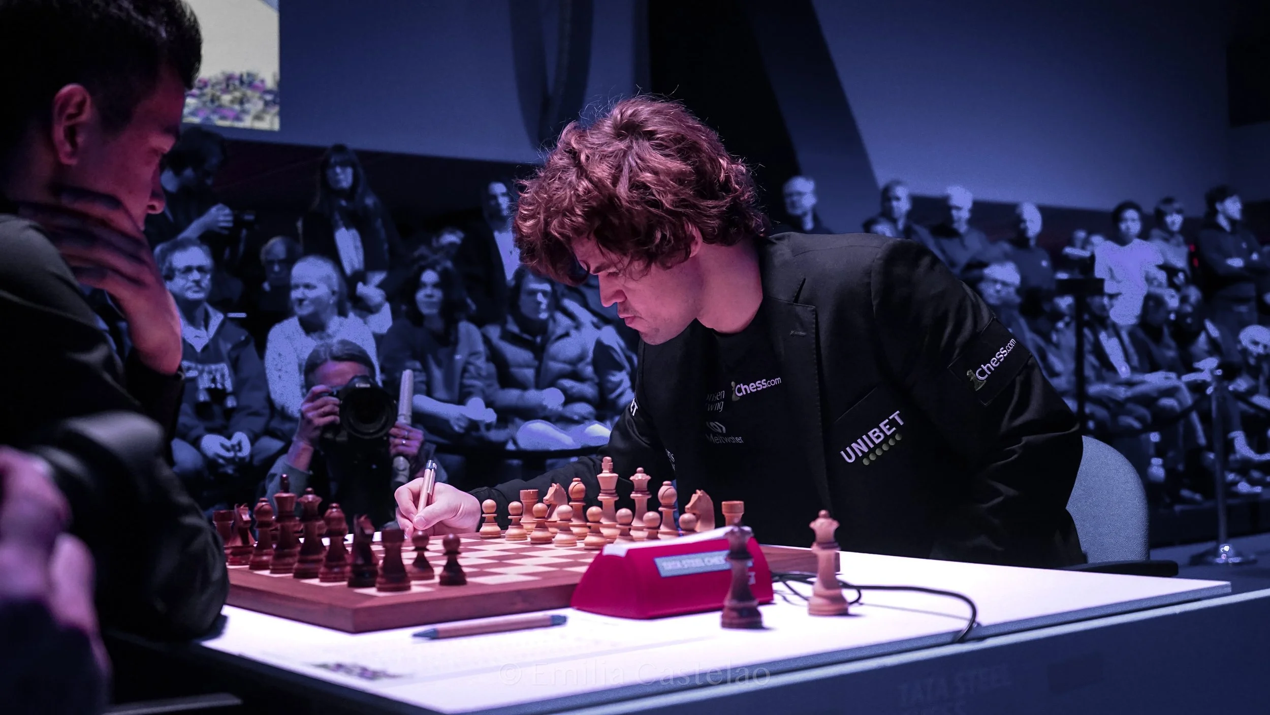 Chess match with one player making a move and the other observing in an audience filled with spectators, some taking photos.