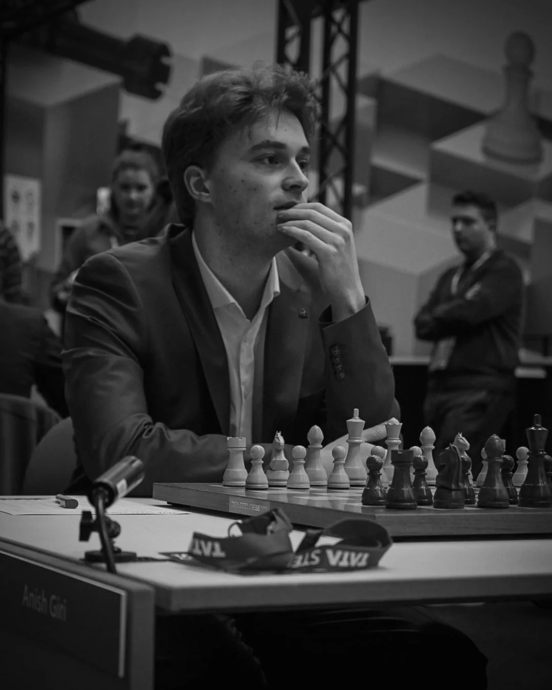 A young man in a suit concentrating on a chess game at a chess tournament, with other people in the background, in black and white.