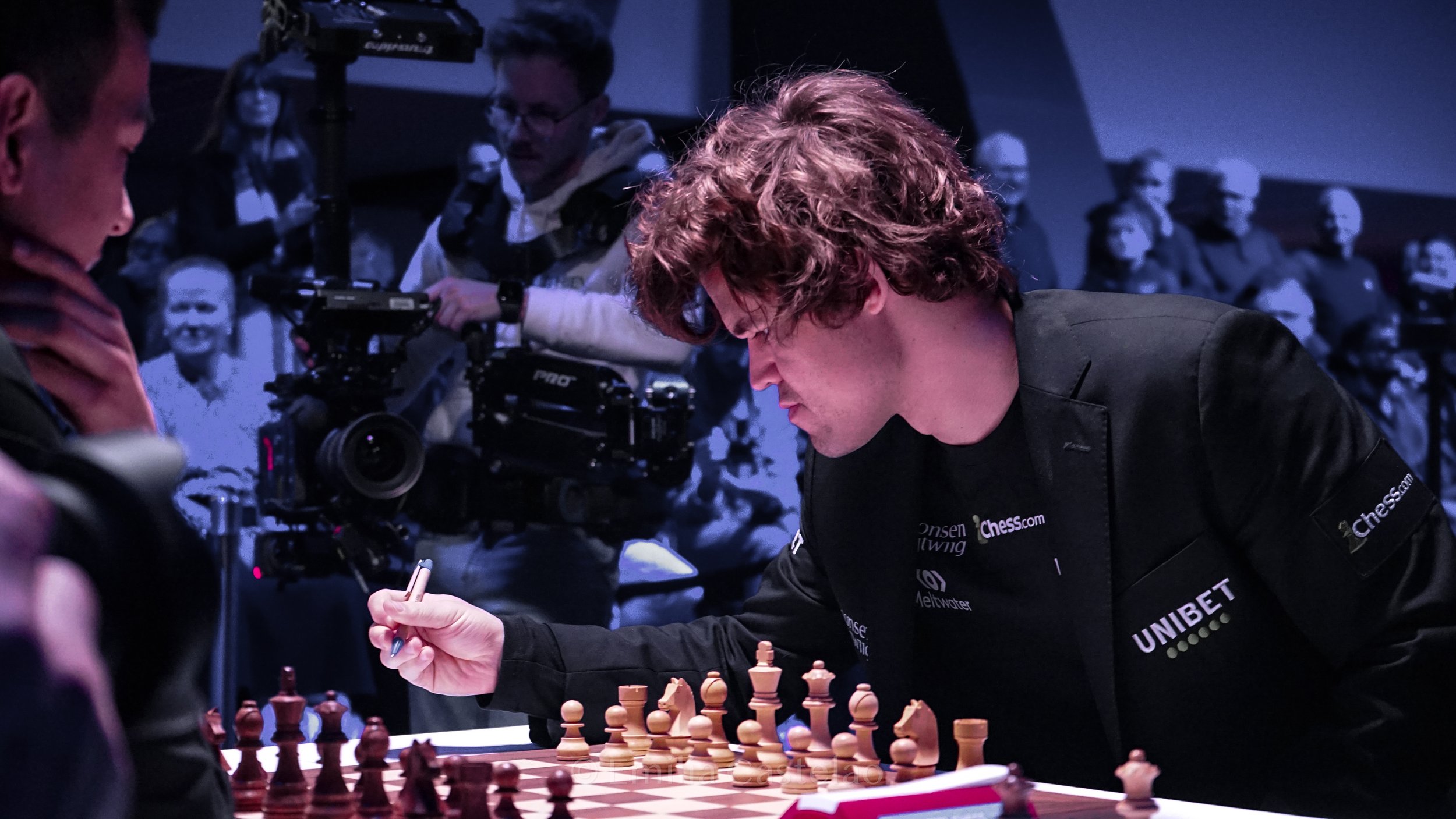 A man with curly hair and glasses making a move in a chess game at a tournament, with a camera crew filming him and a crowd watching in the background.