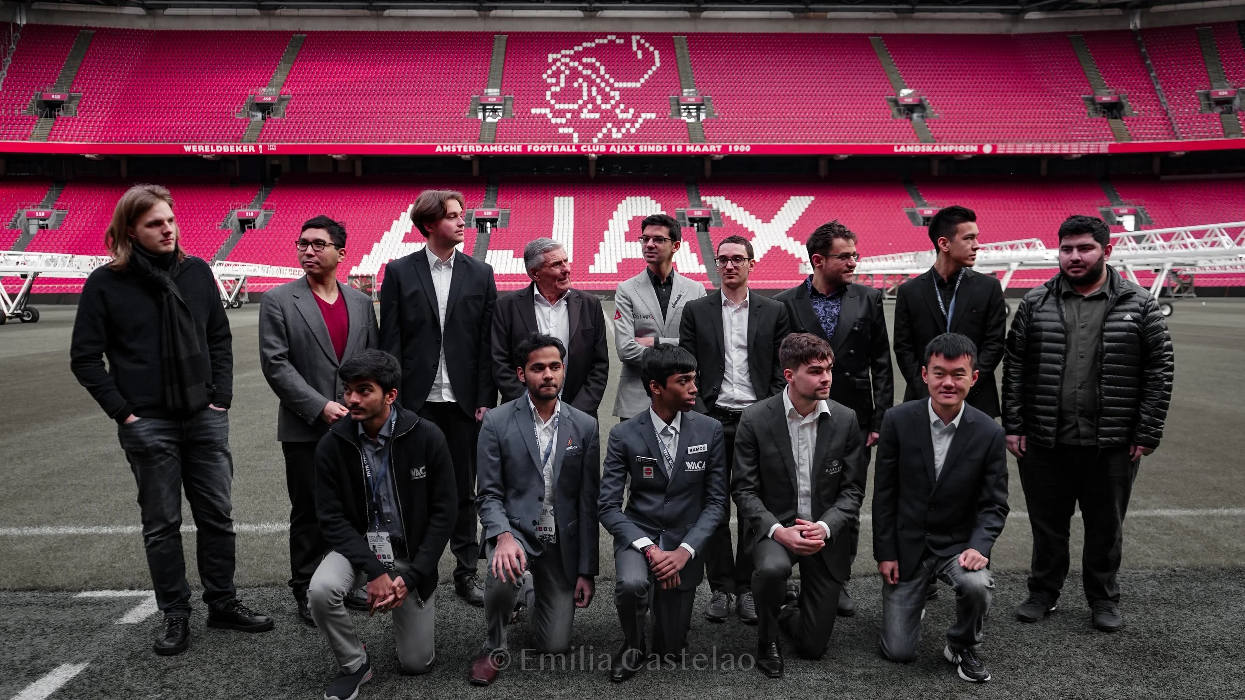 Group of people in business attire posing on a football field inside a stadium with red seats and Amsterdam FC logo in the background.