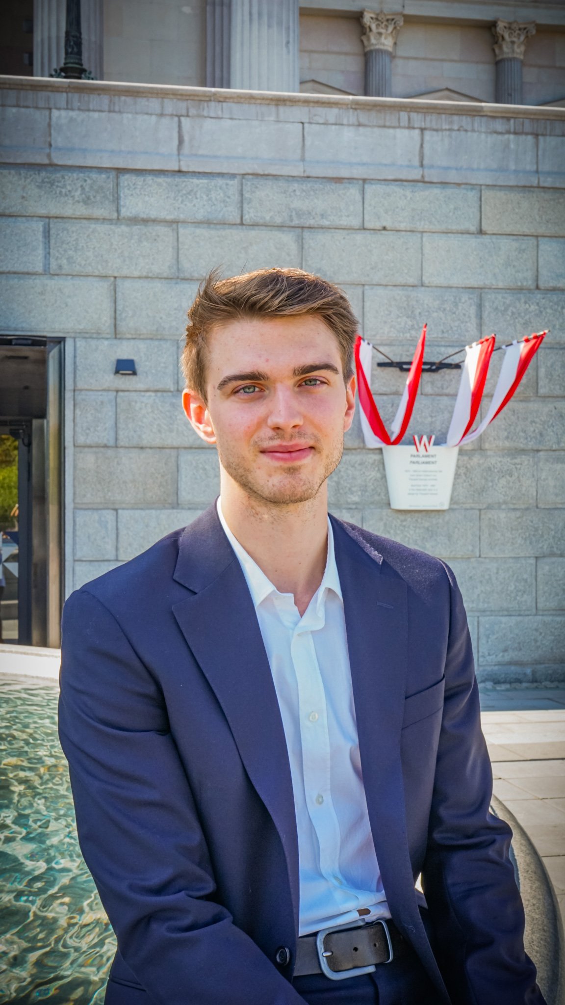 A young man in a navy suit and white shirt sitting outdoors near a fountain, with a stone building and decorative columns in the background.