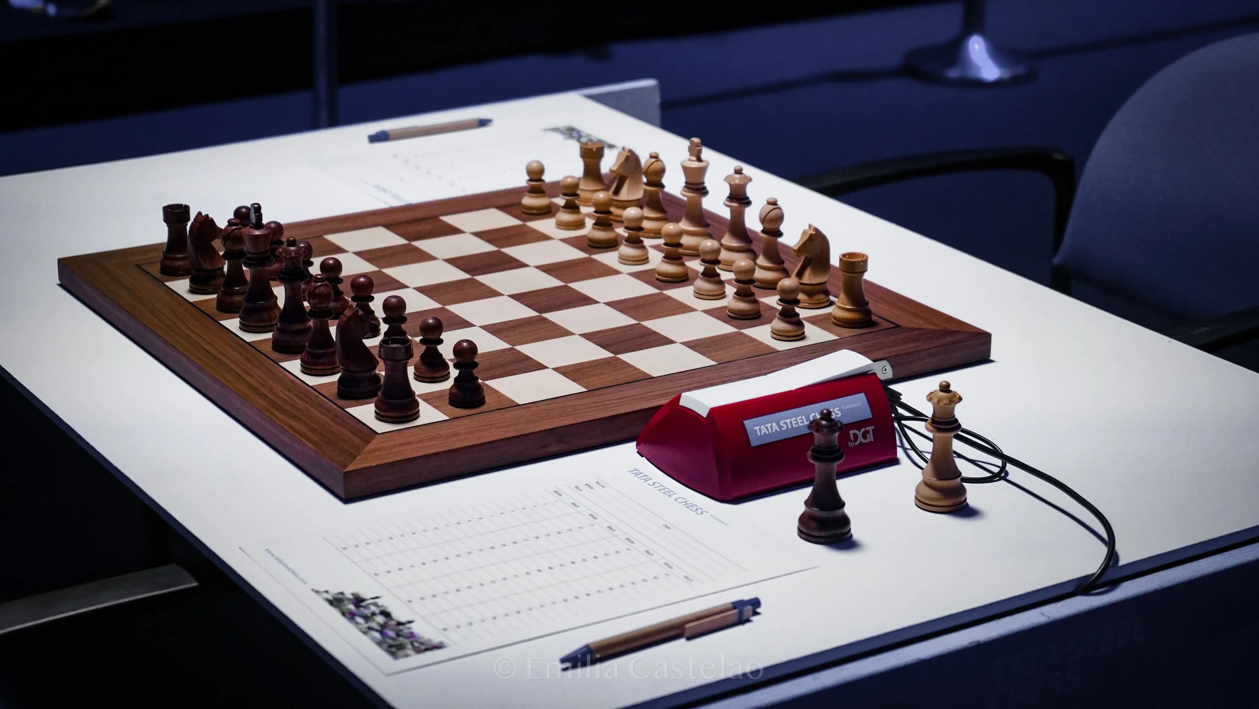 A chessboard set up for a game, with a digital chess clock nearby. The board is wooden with chess pieces in starting positions. There is a pen and a score sheet on the table.