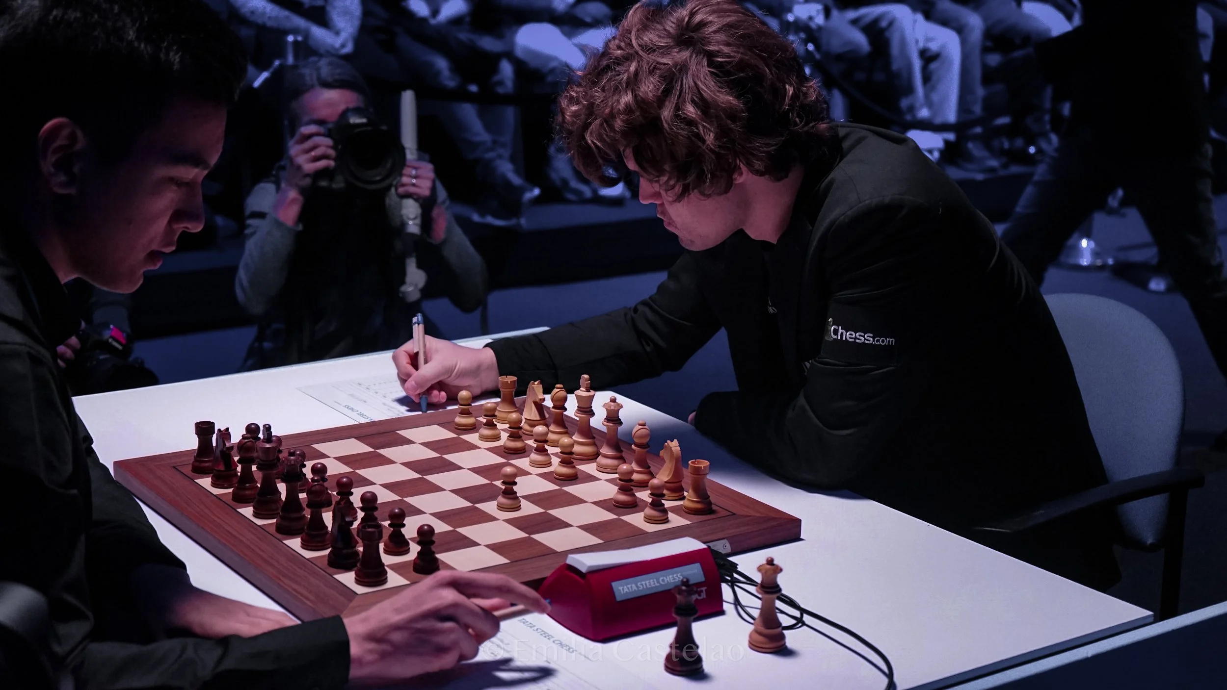 Two men compete in a chess tournament, sitting at a white table with a chessboard, while a woman in the background takes a photograph. The scene is dimly lit, focusing on the players.