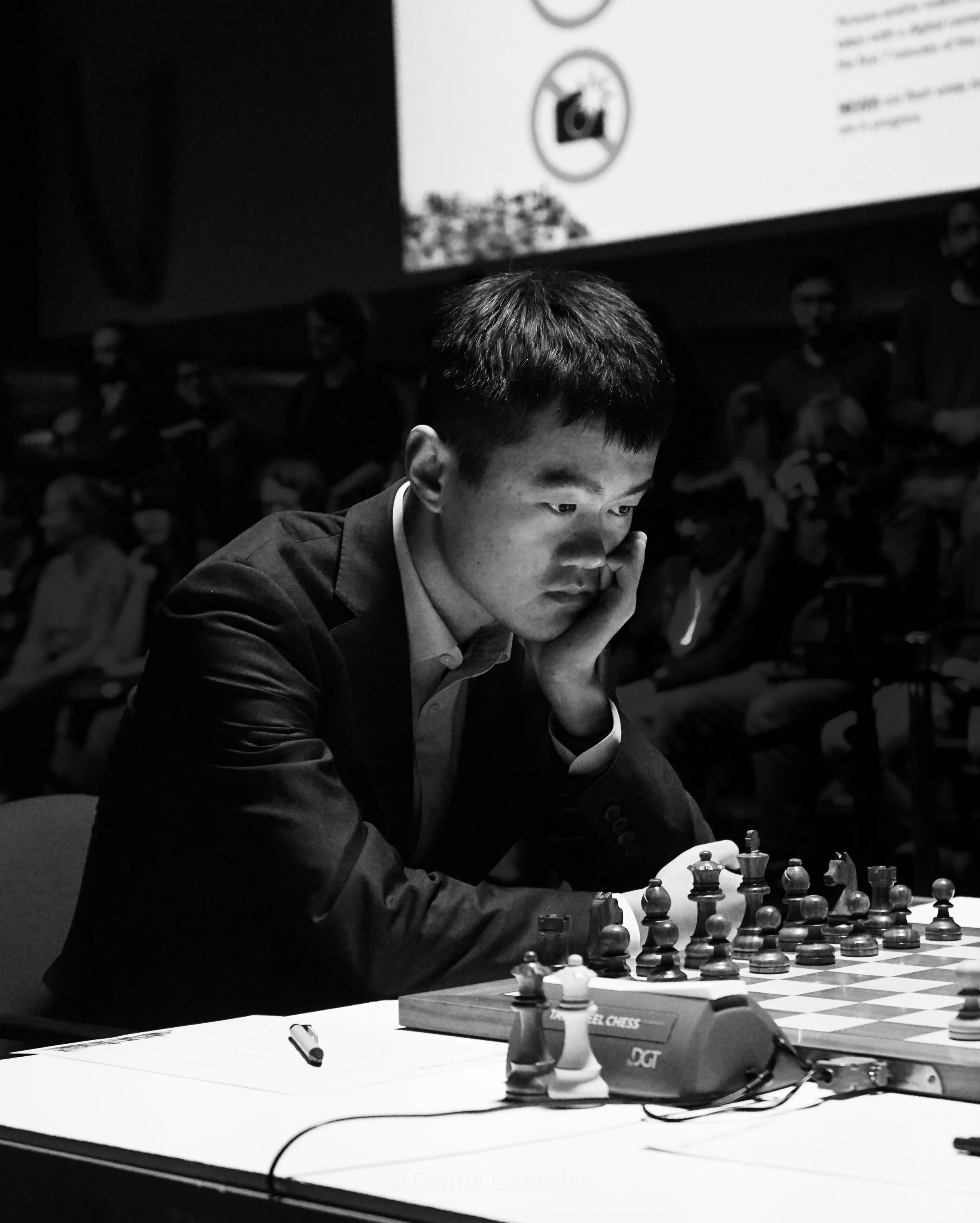 A young man in a suit concentrating on a chess game, seated at a table with chess pieces and a chess clock, with an audience in the background.