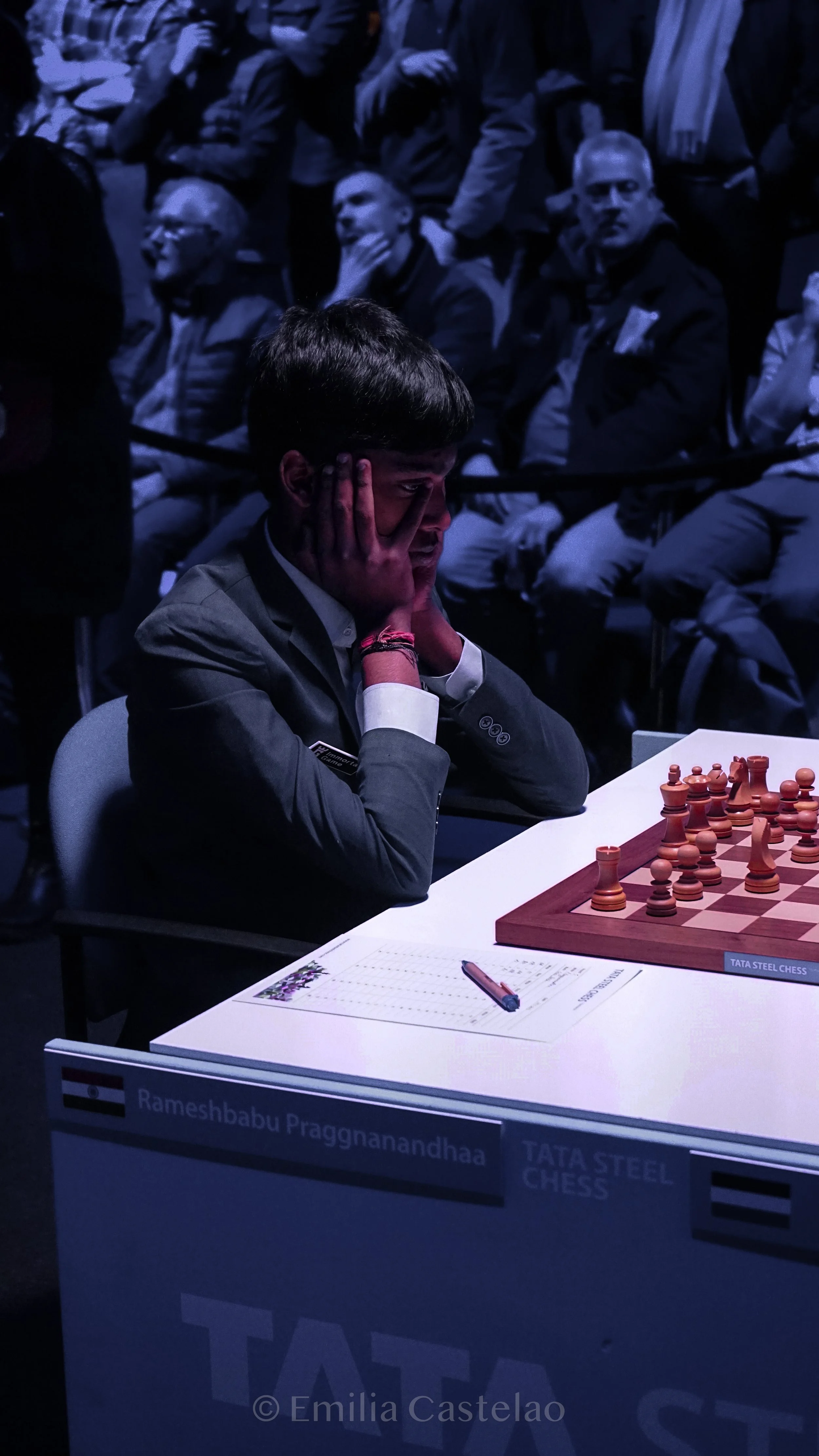 Young man playing chess, surrounded by spectators, in a dimly lit setting.