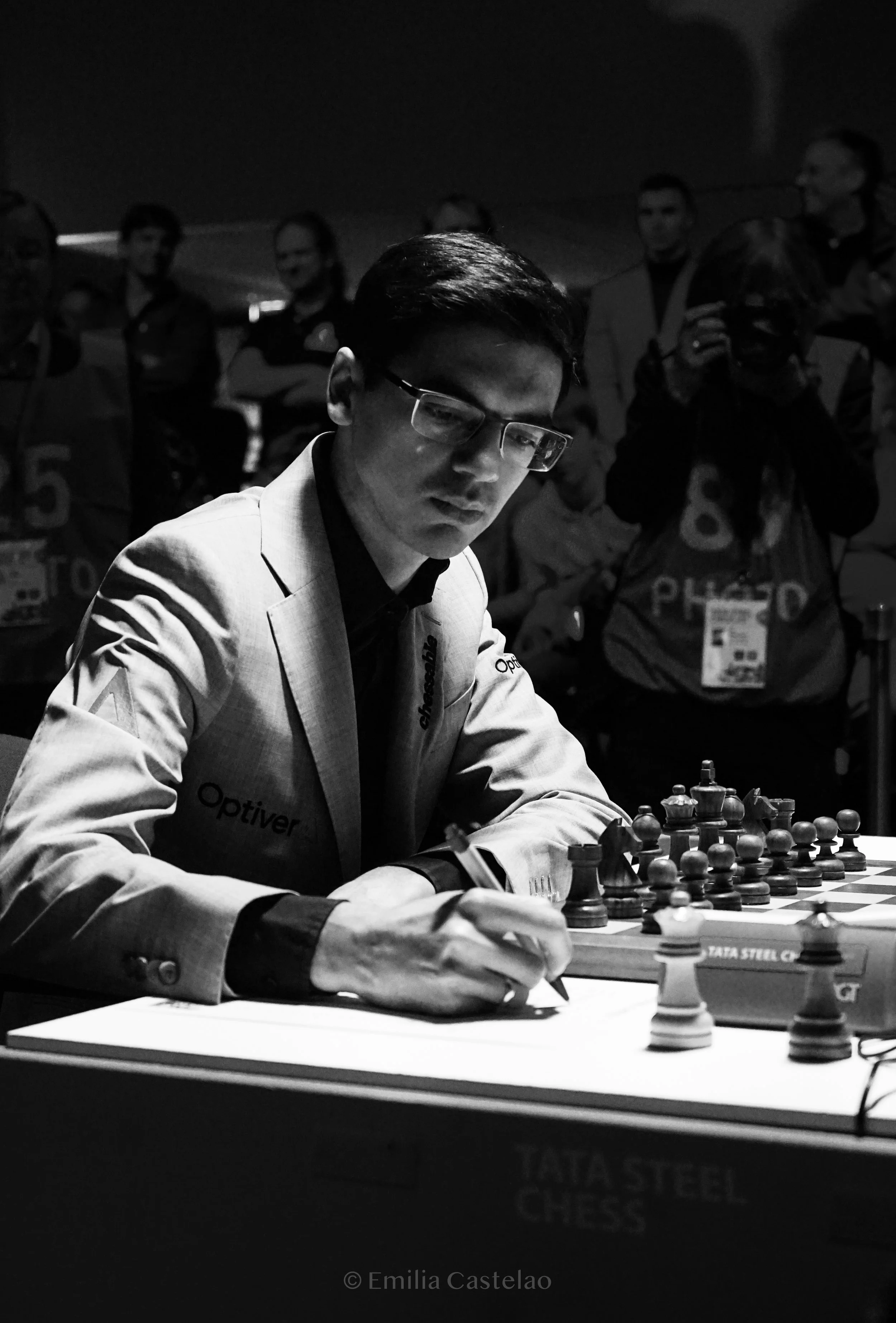 A man wearing glasses and a suit playing chess at a tournament, with spectators watching in the background.