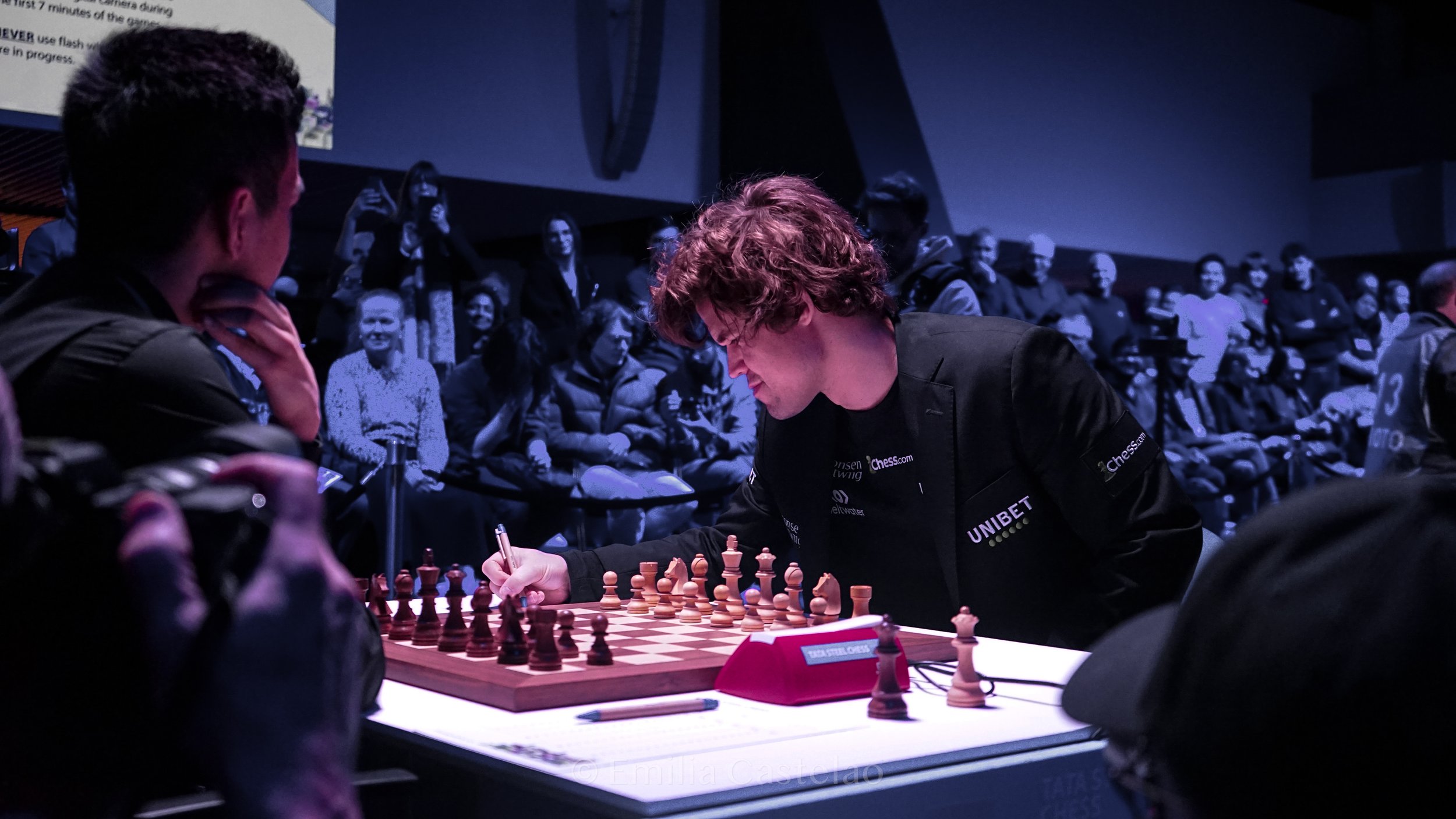 Chess match with a player making a move, surrounded by spectators in a dimly lit room.