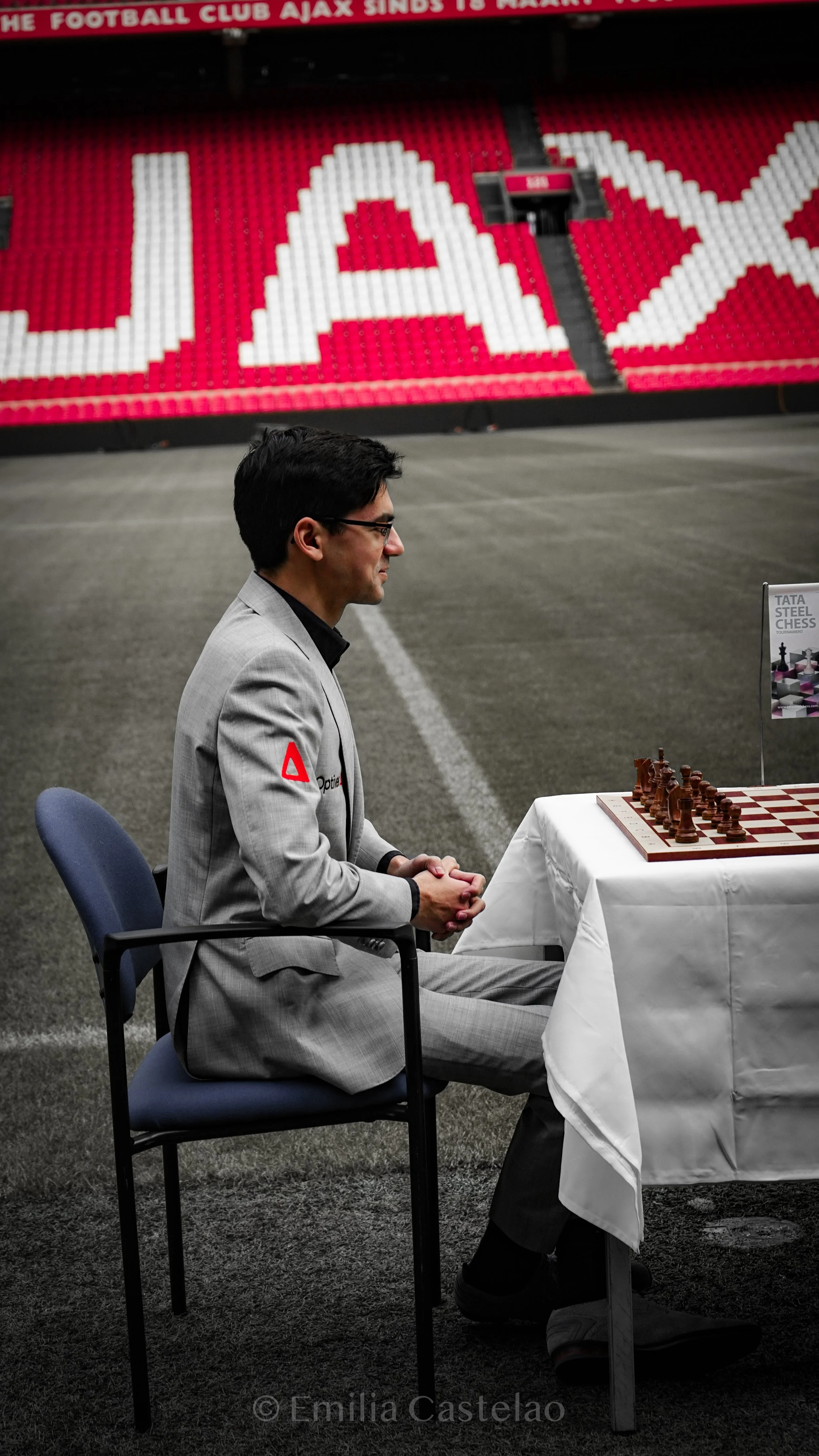 A man in a light gray suit sitting at a table on a gray carpeted indoor sports field, playing chess. Behind him is a large red and white seating area with the letters 'AJAX' spelled out in lego-like blocks.