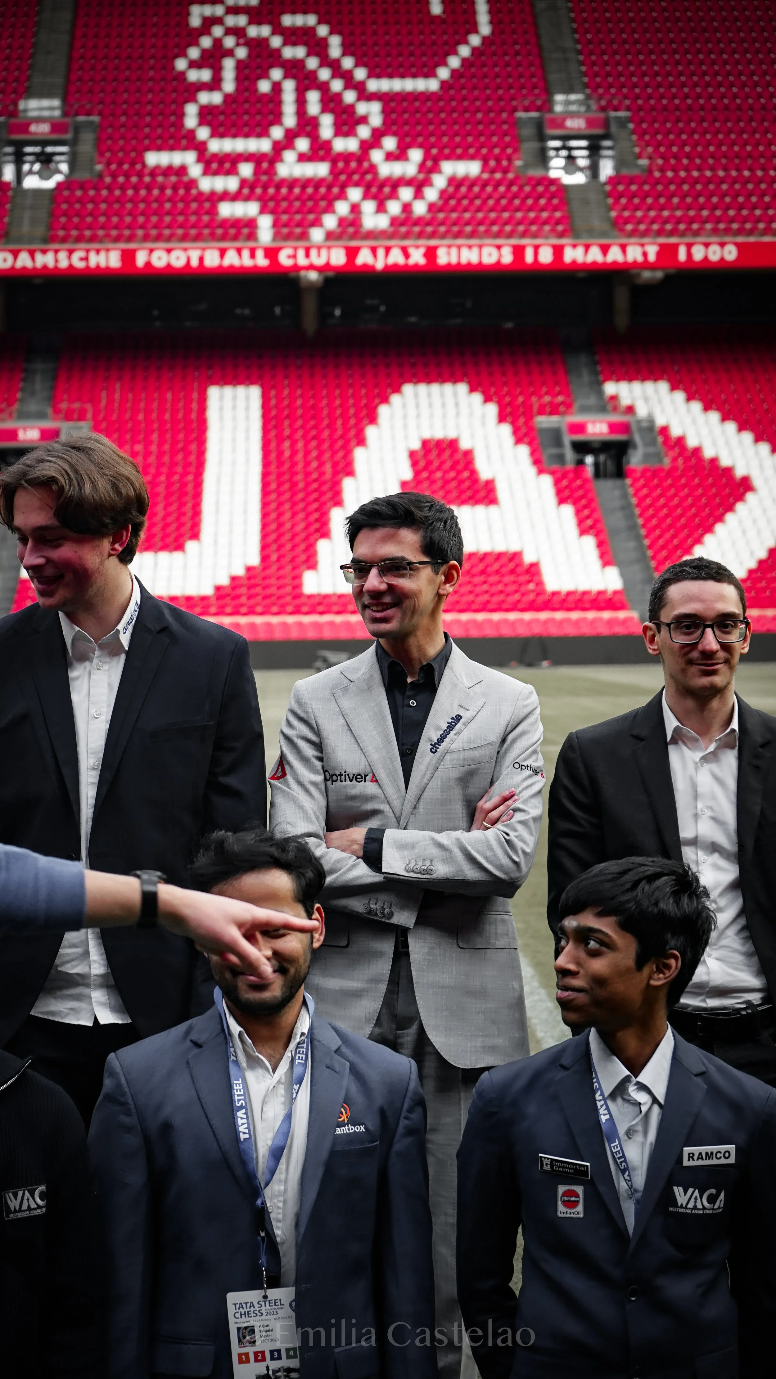 Group of men in suits standing on a football field inside a stadium with red and white seating, some with logos and patterns, and a large digital clock.