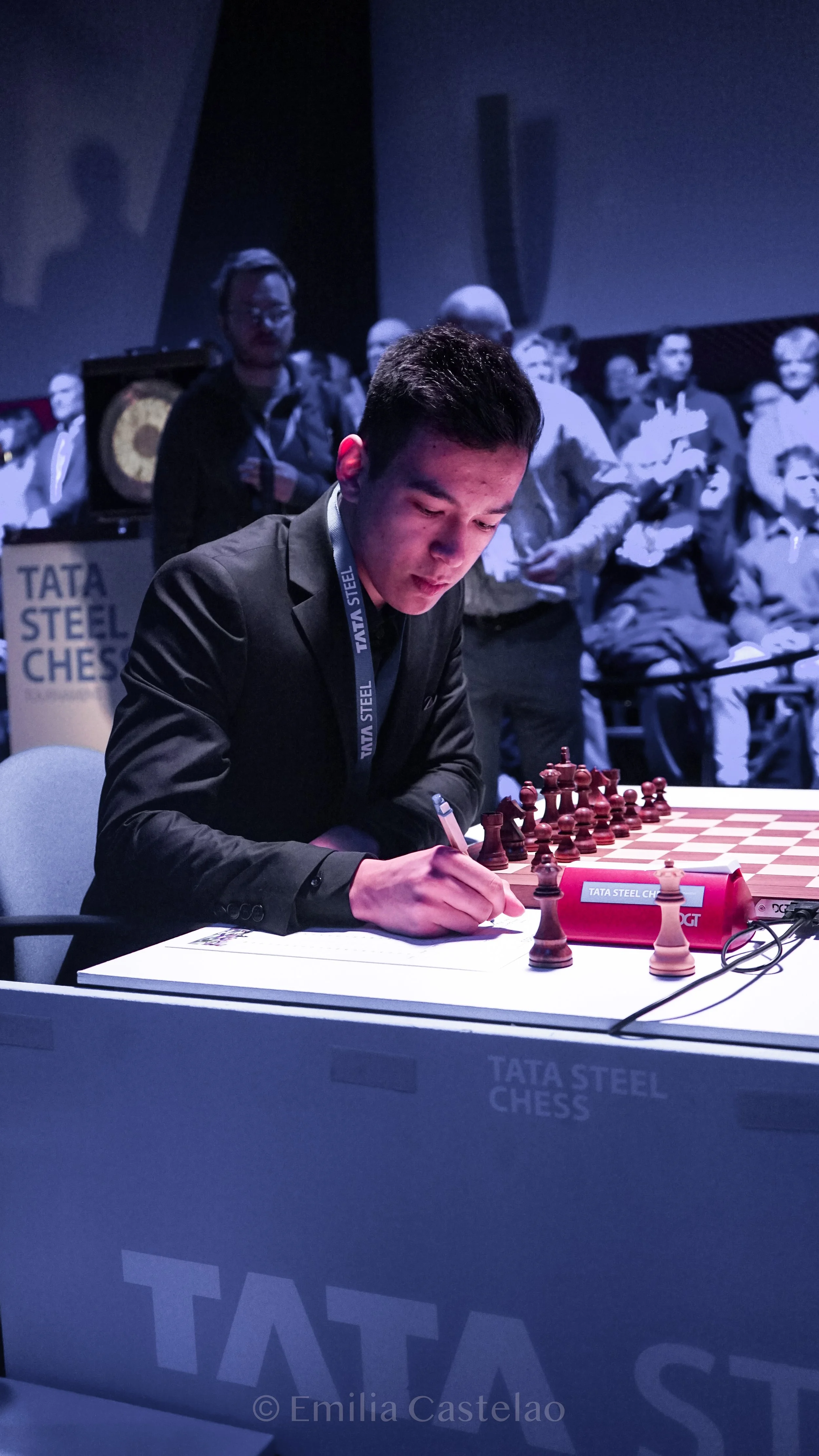 A young man writing at a chess table during a chess tournament with an audience in the background.