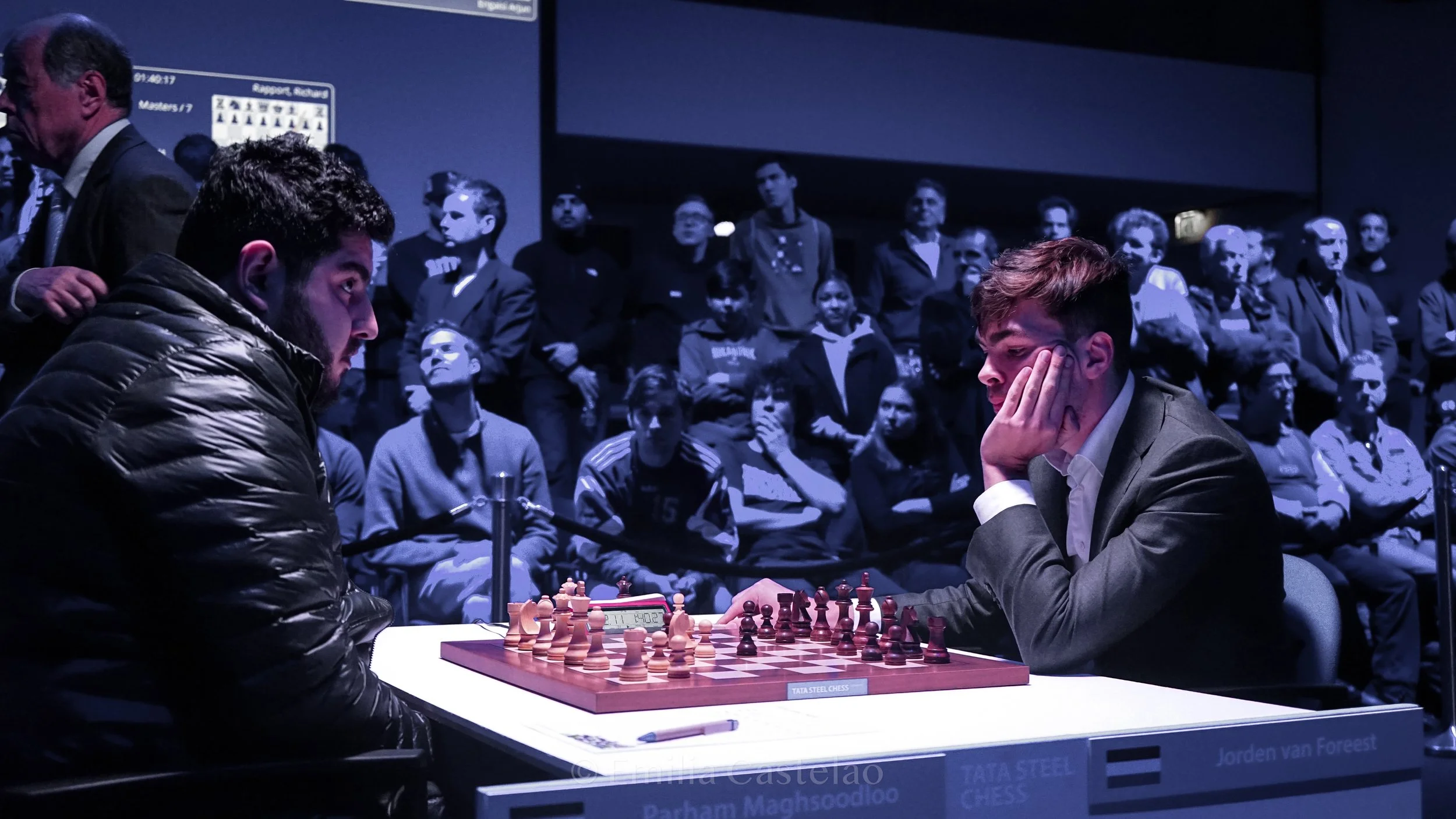 Two young men playing chess in a competitive match, with a digital clock on the table, and an audience watching behind them in a dark room.
