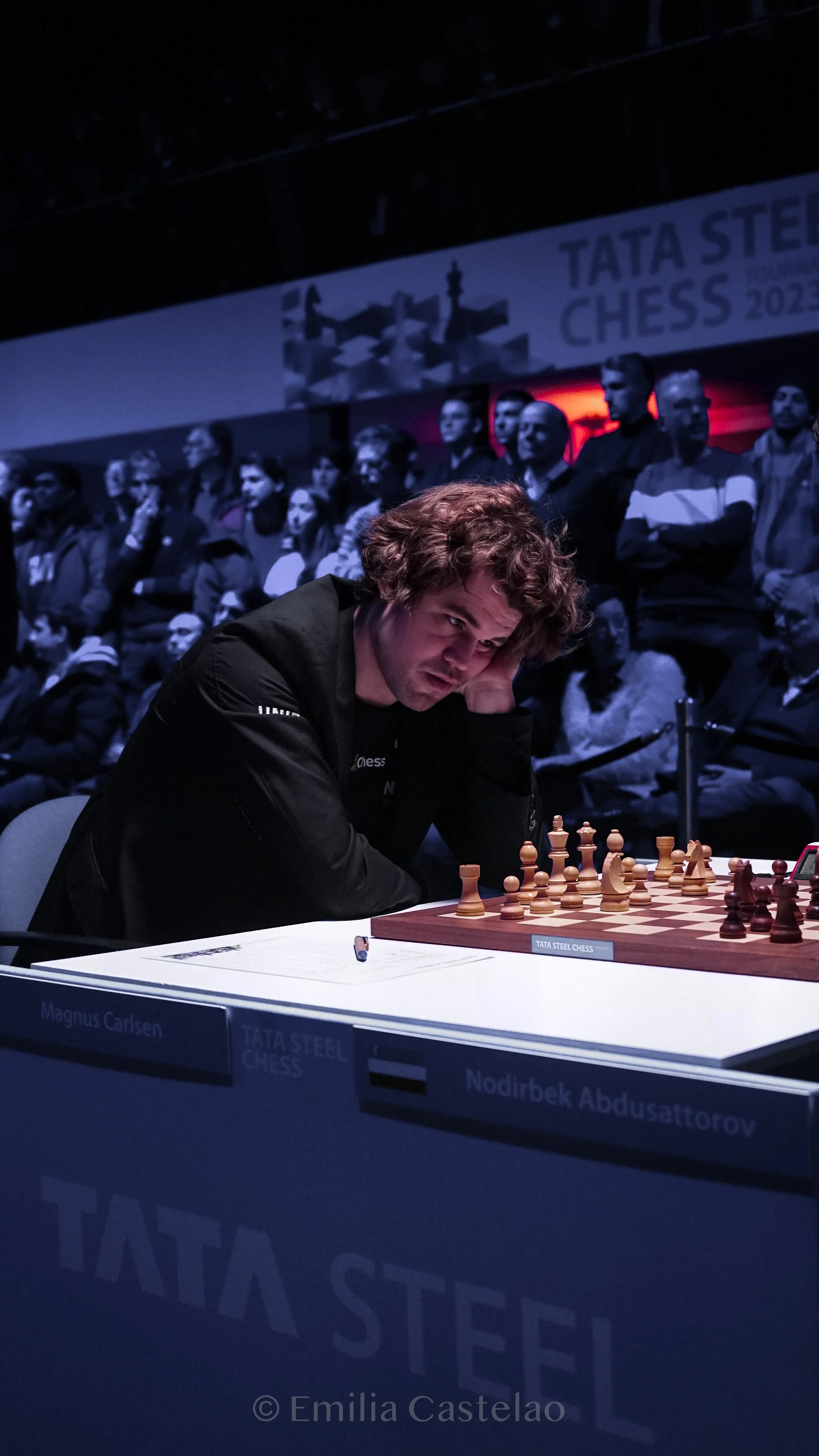 Chess player with curly hair resting his head on his hand during a match at the Tata Steel Chess tournament, with a crowd observing in the background.