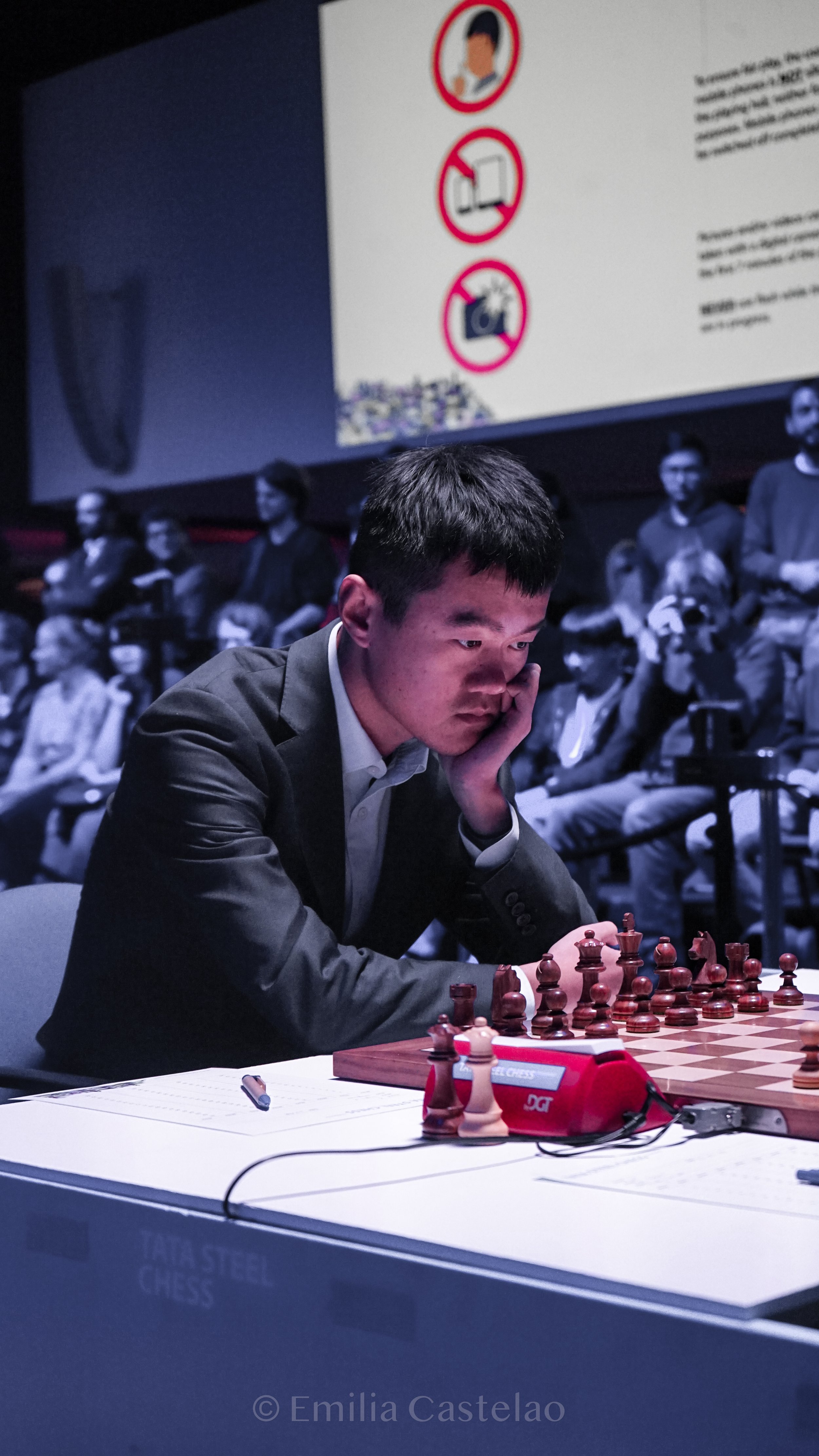 A young man intensely concentrating on a chess game at a tournament, sitting at a table with a chessboard, timer, and scoresheet, with spectators in the background and a large screen displaying rules.
