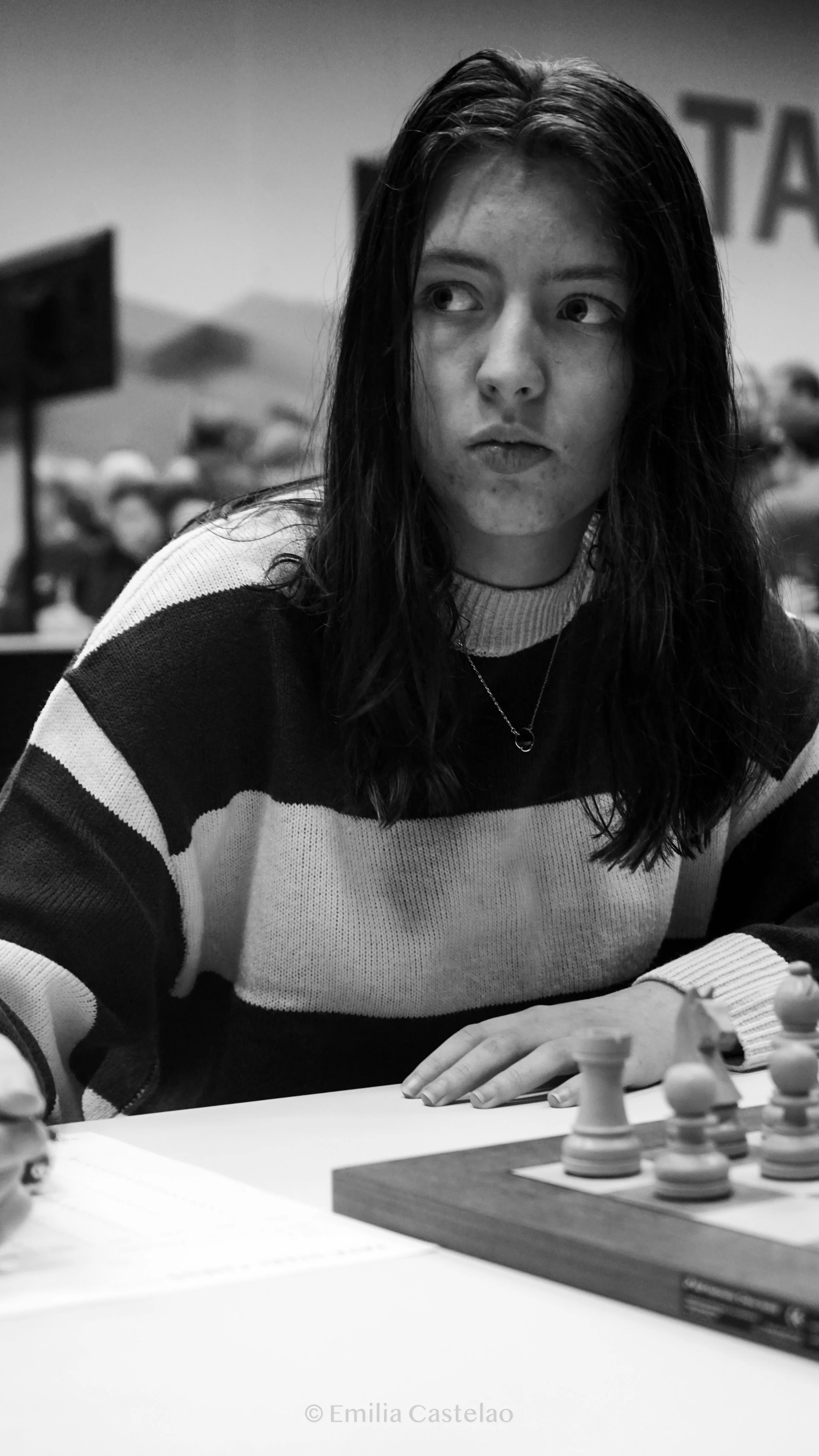 A young woman with loose dark hair, wearing a striped sweater, sitting at a table with a chessboard, looking to the side with a contemplative expression in a casual indoor setting.