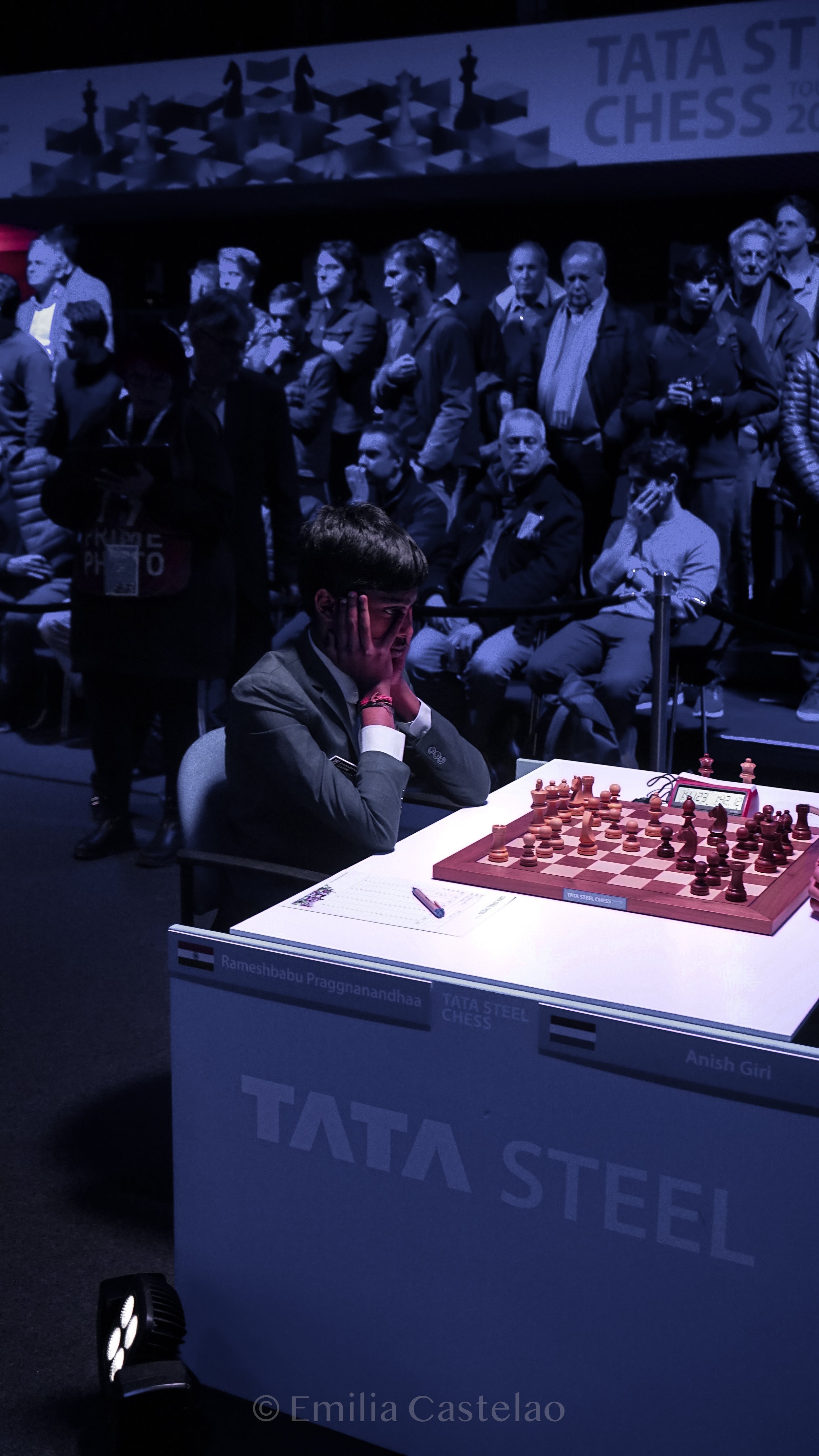 A young boy deeply concentrating on a chess game at the Tata Steel Chess tournament, with a crowd of spectators watching behind him.