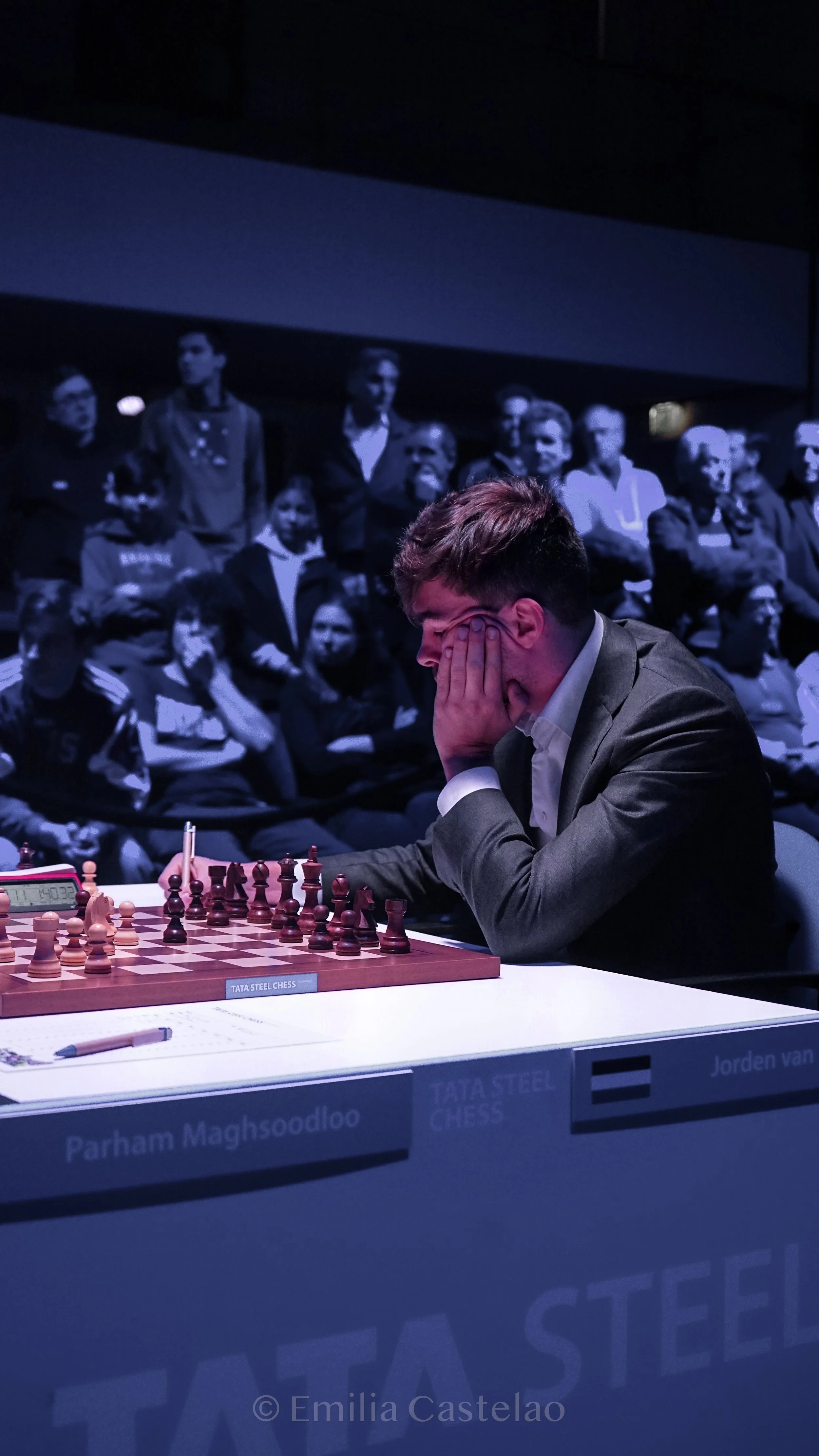 A young man in a suit is playing chess during a tournament, sitting at a table with a chessboard. He has his hands on his face, focused on the game. In the background, a large screen displays a crowd of spectators. There is a sign on the front of the