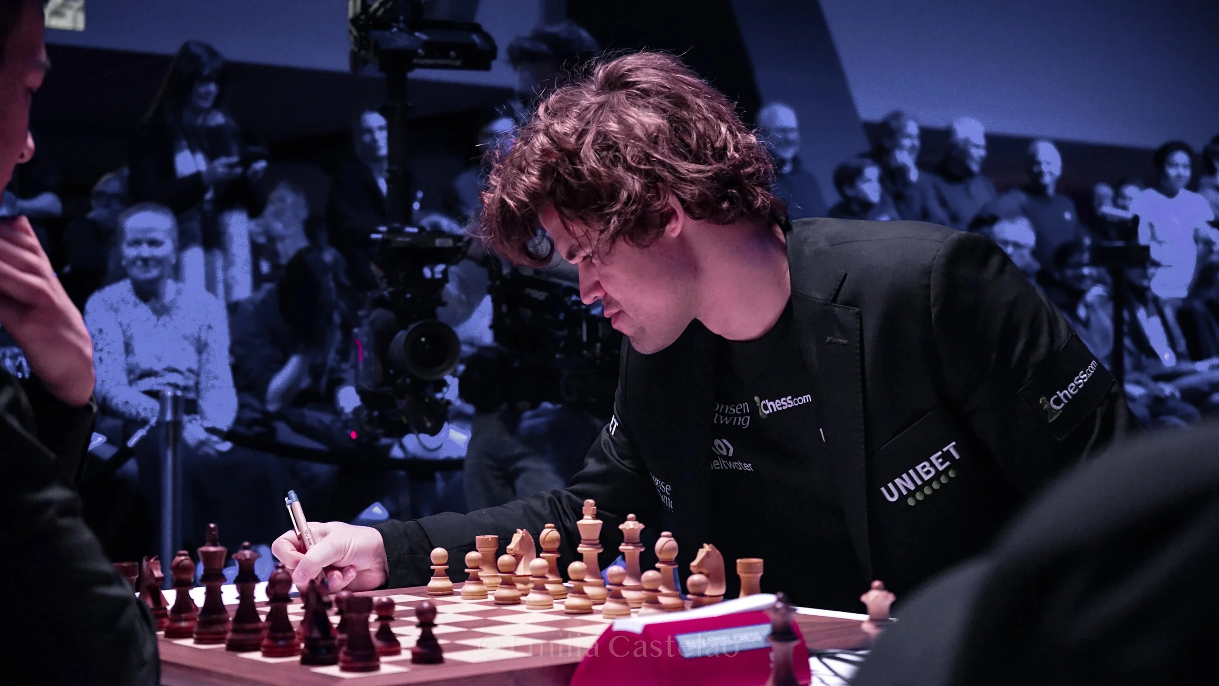 A man with curly brown hair and glasses, wearing a black jacket with various sponsor logos, is playing chess at a tournament. He is making a move on the chessboard while others and camera operators observe in the background.
