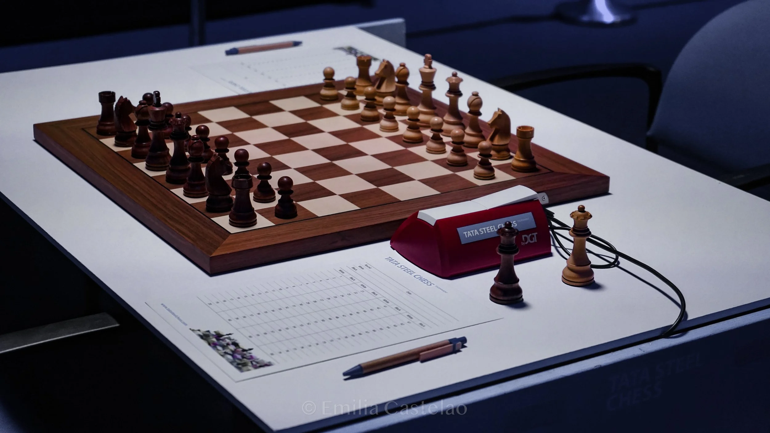 A chessboard with wooden pieces set up for a game, placed on a white table. A clock and score sheet are nearby. The scene appears to be in a tournament setting.