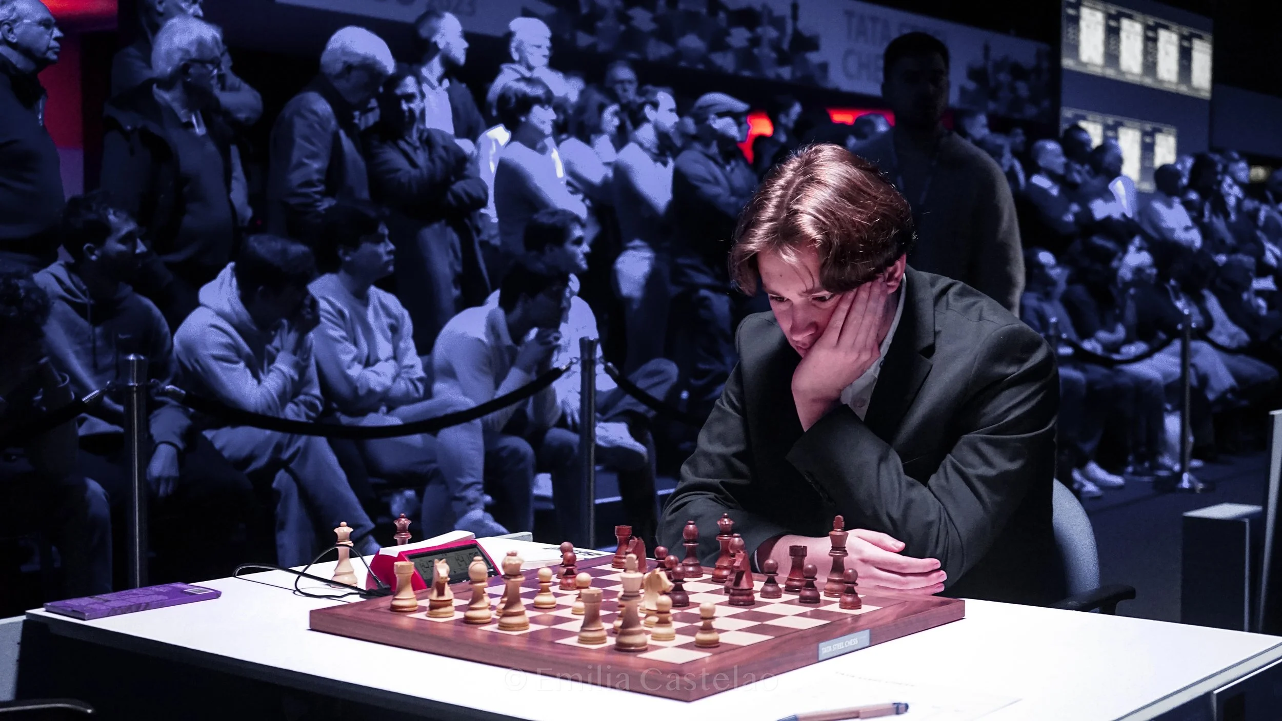 A young man in a black suit playing chess at a tournament table with a crowd of spectators watching in the background.