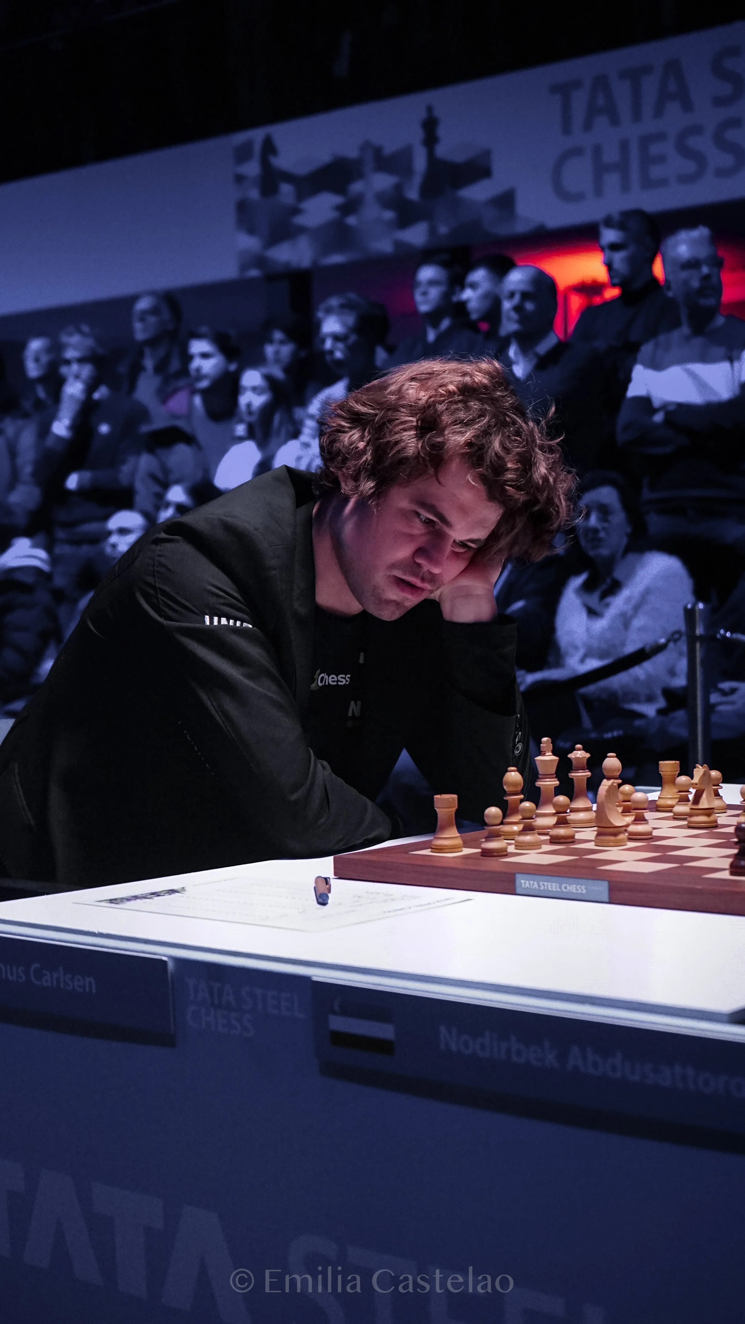 A young man playing chess at a tournament, surrounded by spectators, with a dark background and overhead lighting.