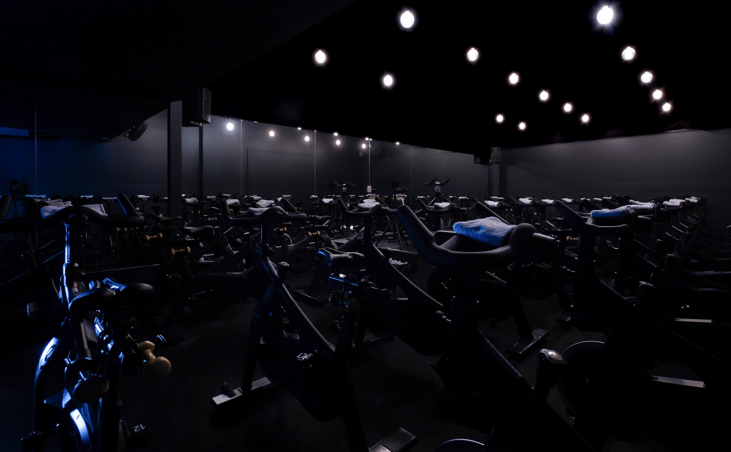 A dimly lit indoor cycling studio with multiple stationary bikes lined up in rows, each equipped with towels and screens, illuminated by a series of overhead lights.