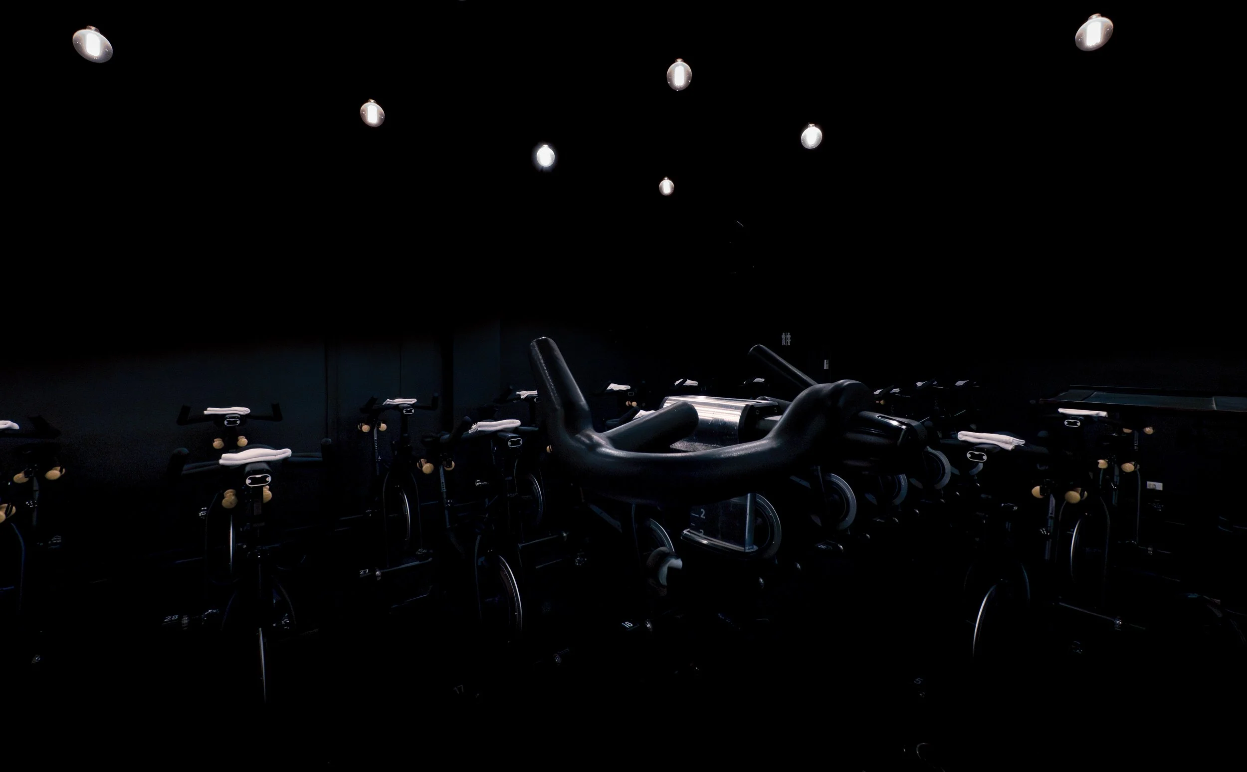 Indoor cycling studio with multiple stationary bikes and a large fitness wheel in front, dimly lit with black walls and ceiling lights.