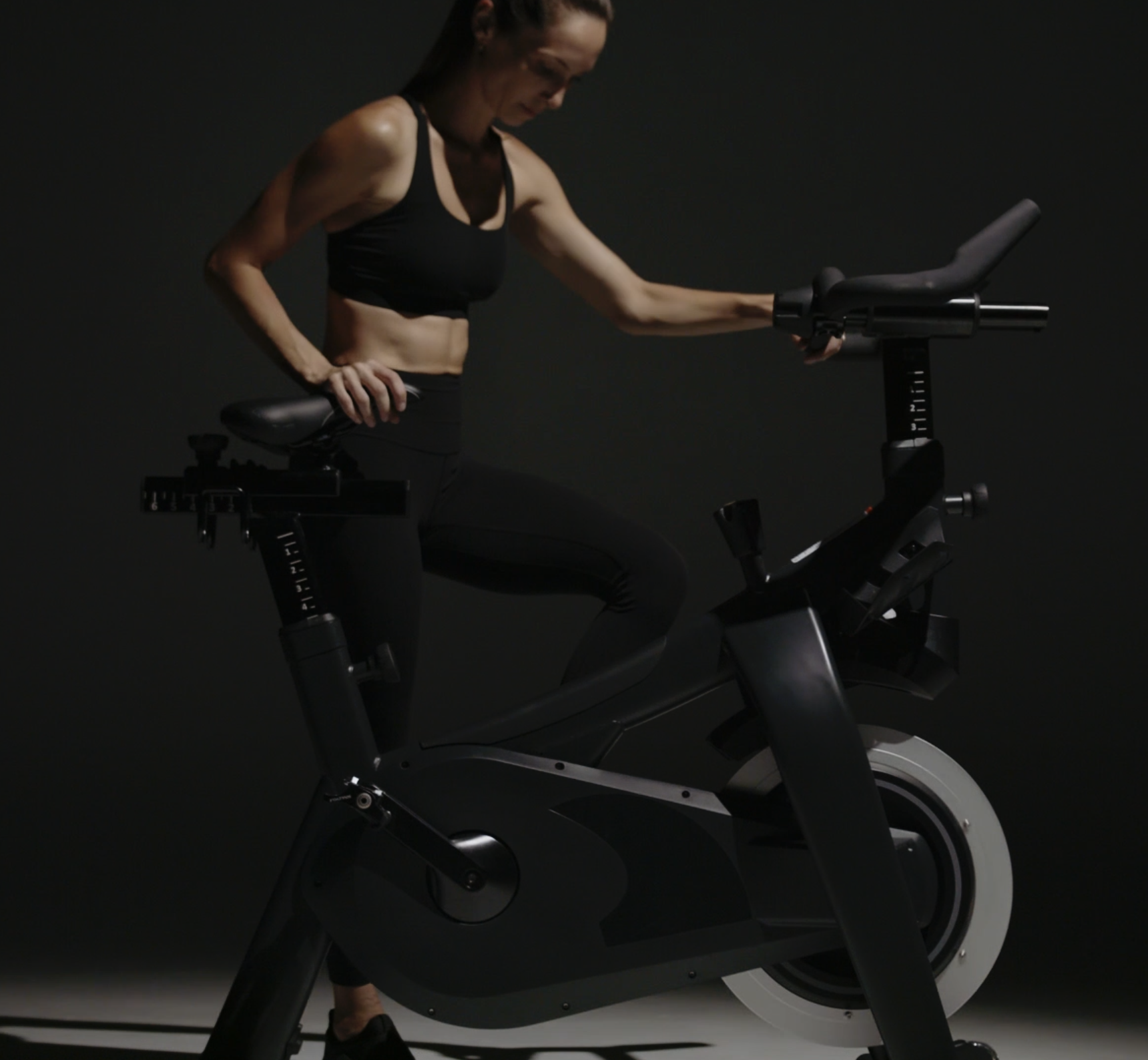 A woman in athletic clothing is riding a stationary exercise bike in a dark room with minimal lighting.