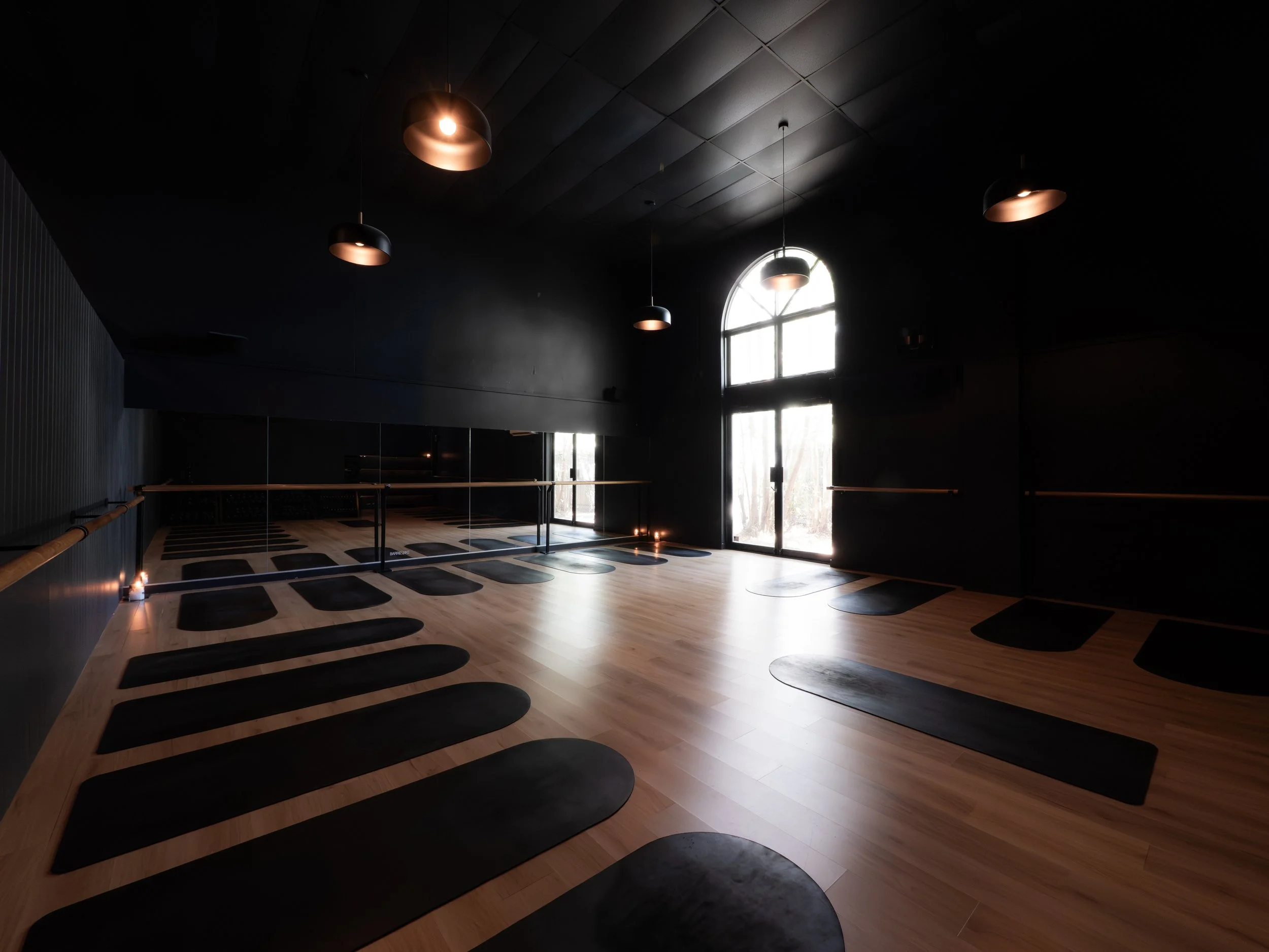 Empty yoga or pilates studio with black mats arranged in rows on a wooden floor, black wall, large window with natural light, and black pendant lights hanging from ceiling.