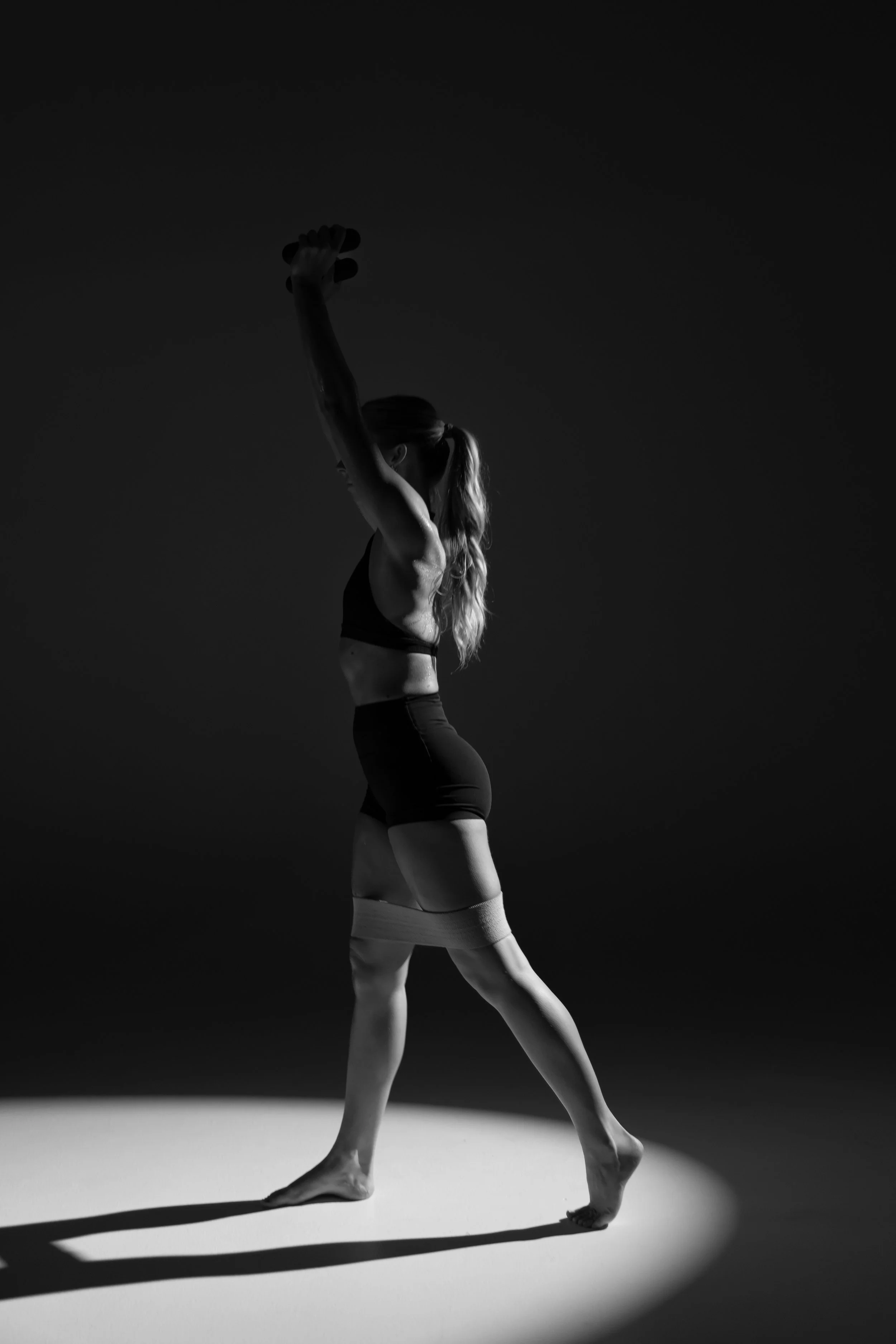 Black and white photo of a woman in a dance pose. She is standing on one leg, with the other leg slightly bent, and her arms raised above her head. Her hair is pulled back in a ponytail. The background is dark and contrasts with the light surface she is standing on, casting a shadow.
