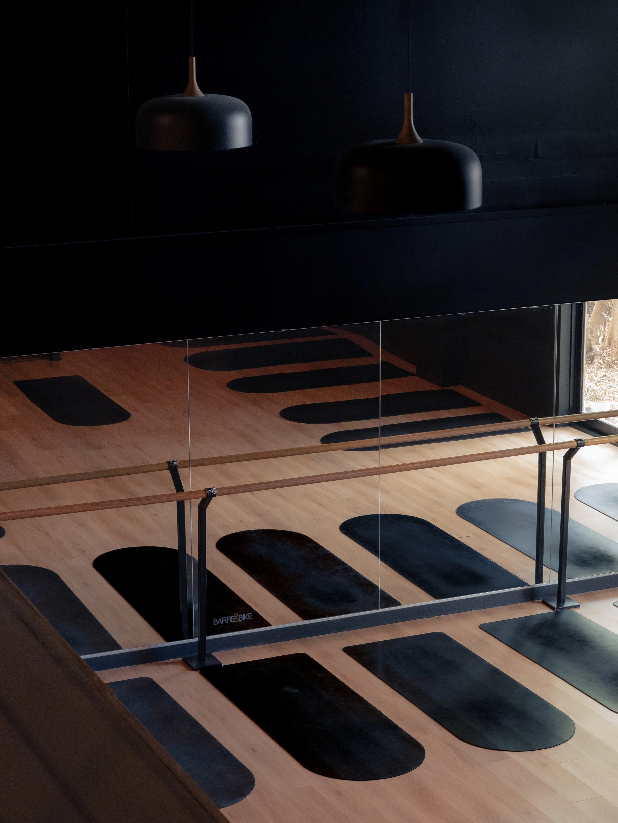 A yoga or exercise studio featuring black mats on a wooden floor, a black wall with hanging black and brown lamps, a mirrored wall reflecting the space, and a railing with vertical bars and wooden handrails.