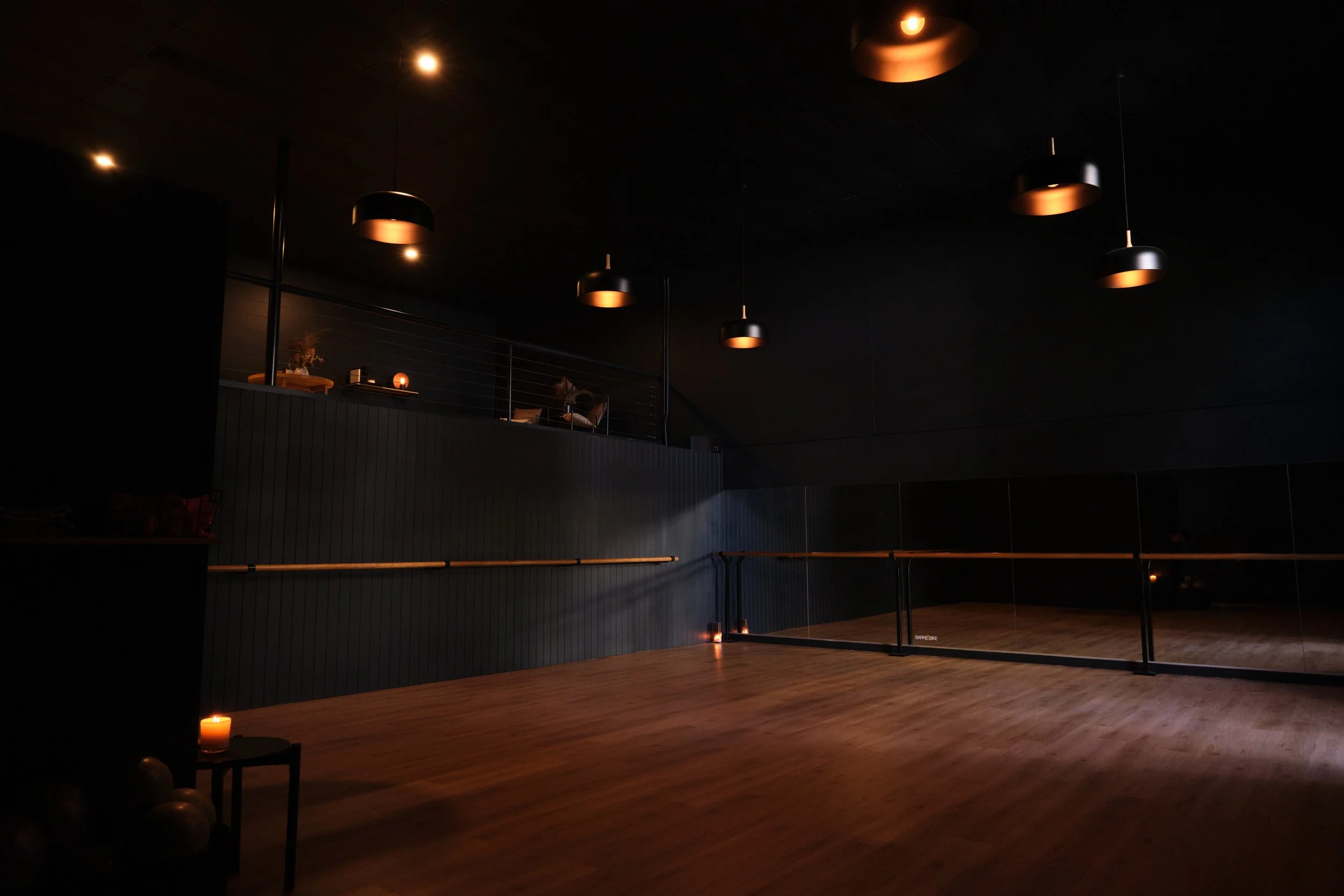 An empty room with wooden flooring and black walls, featuring several modern black pendant lights hanging from the ceiling, a small table with candles in the foreground, and a balcony with chairs and plants in the background.