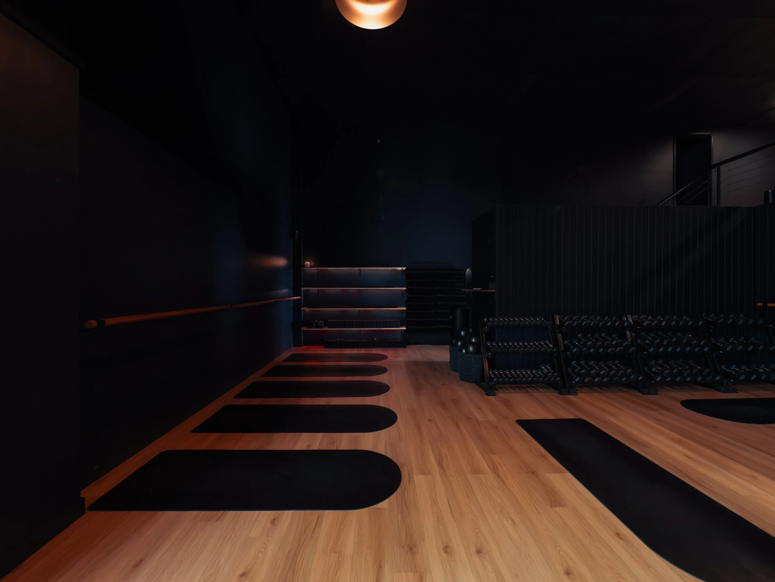 A dark gym room with exercise mats placed on wooden floor, a rack of dumbbells, and some foam rollers. The room has black and dark grey walls with wooden accents and minimal lighting.