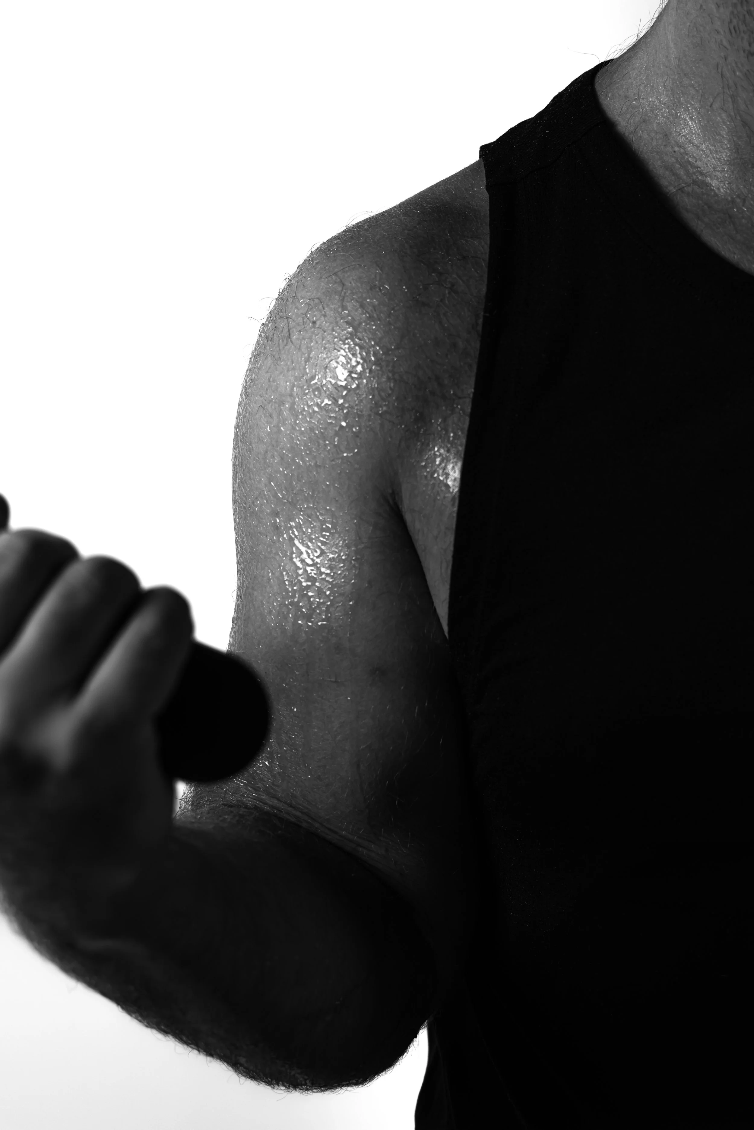 Close-up black and white photo of a person's shoulder, upper arm, and part of an arm with visible skin texture and small hair, wearing a black sleeveless top, with fingers holding an object.