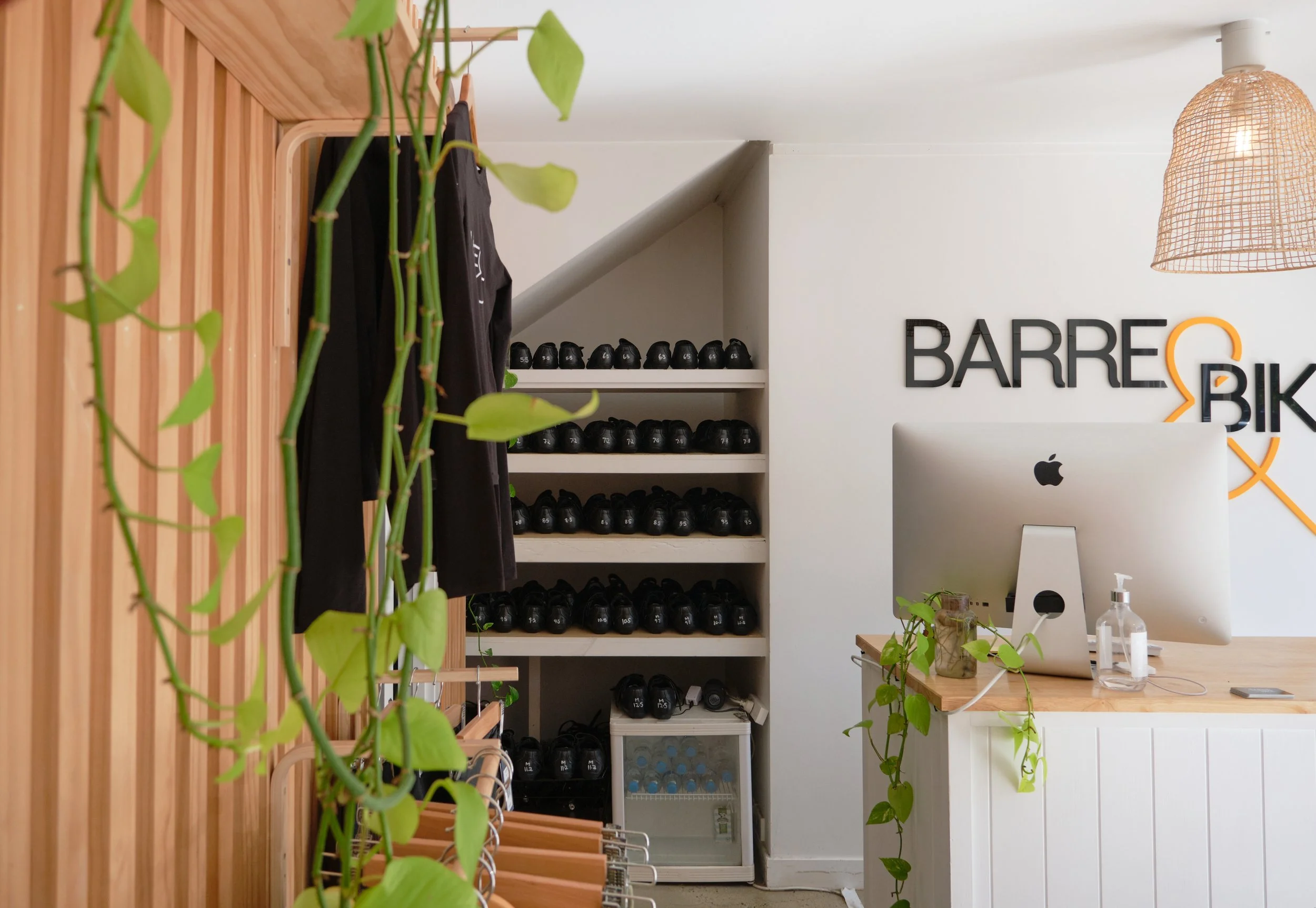 Interior of a fitness or wellness studio with a wall display of black kettlebells arranged by size in a corner, a white reception desk with a computer monitor, a wooden countertop, and a hanging woven light fixture. Green leafy plants are visible in the foreground and a sign on the wall reads 'Barre & Bik'.