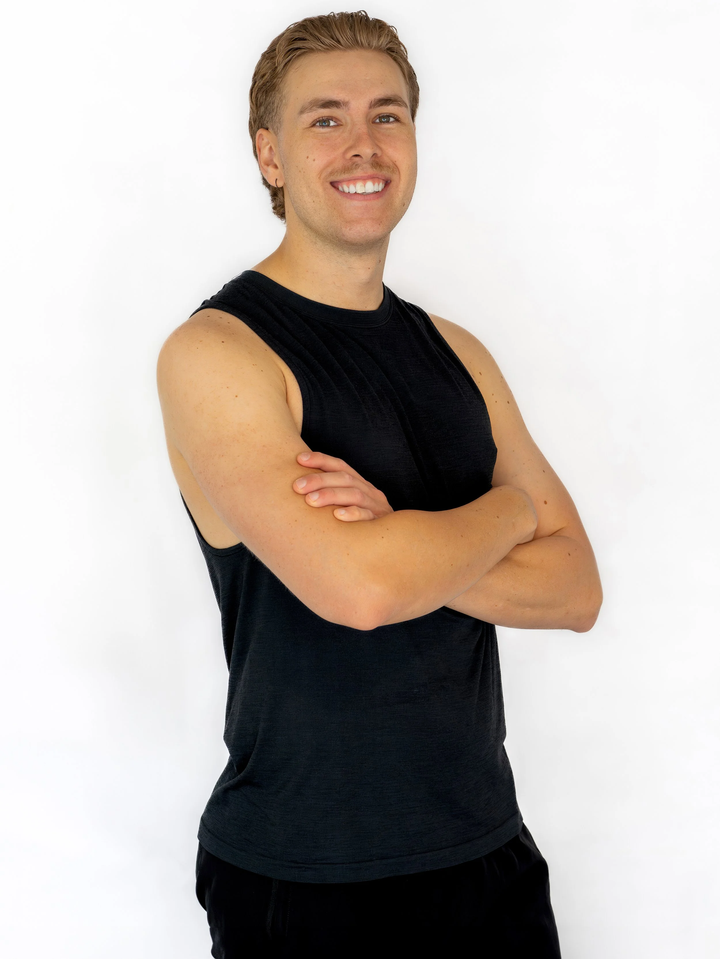 A smiling young man with painted arm muscles, wearing a black tank top and black shorts, standing against a white background.